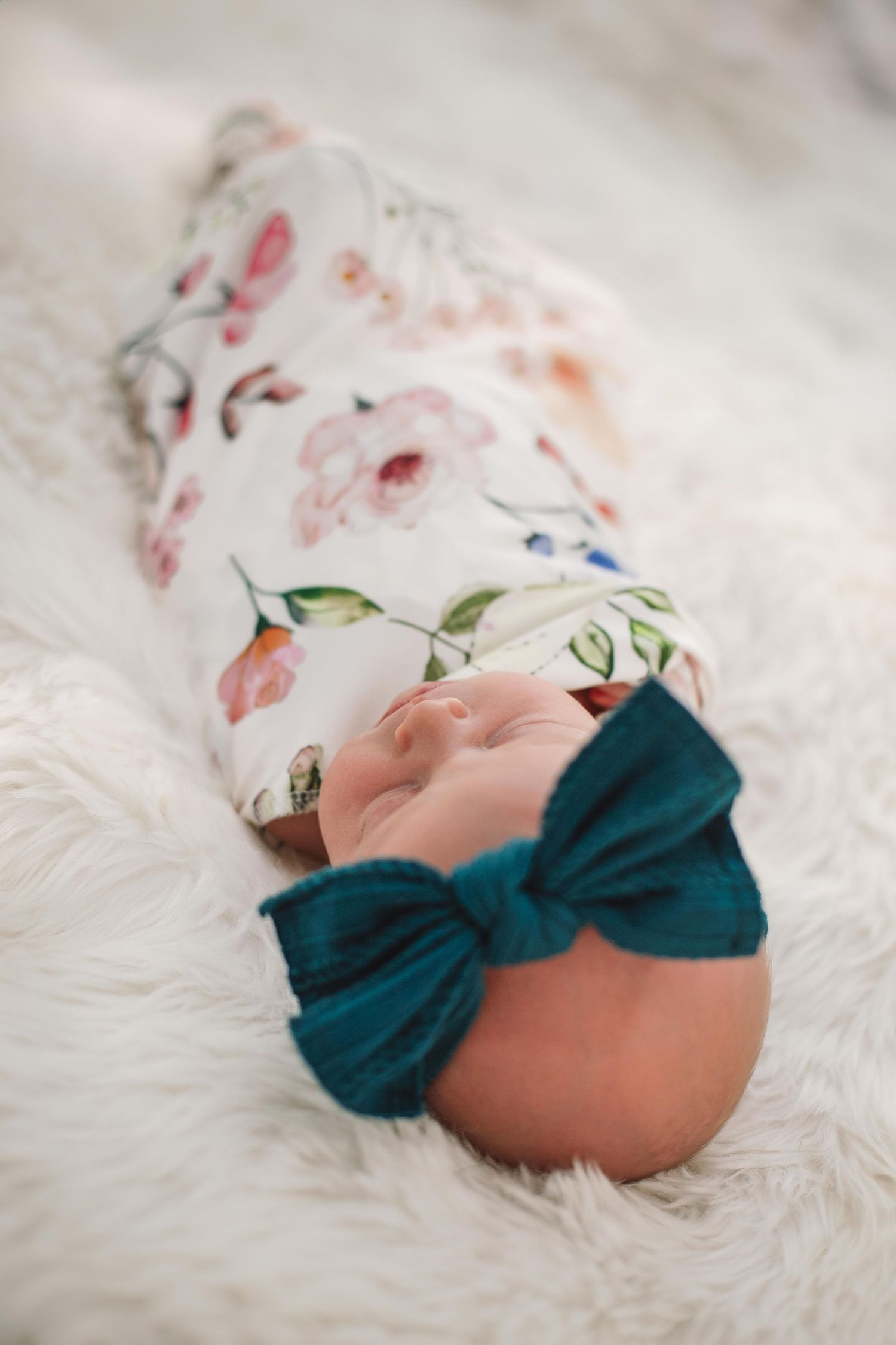 Baby in teal bow headband and floral print sleeps on soft white fur.