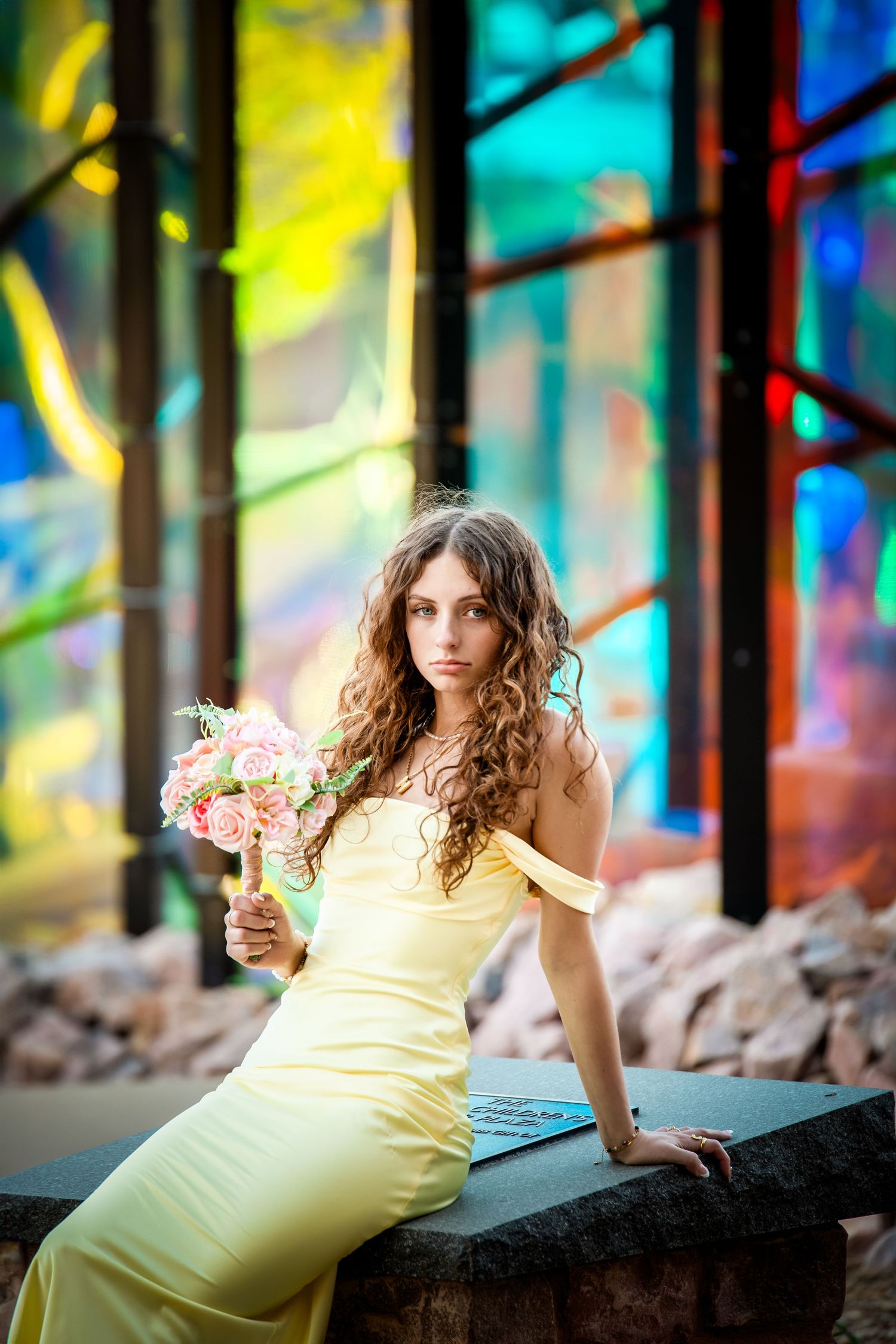 A woman in a yellow dress poses gracefully against colorful stained glass windows while holding a floral bouquet.