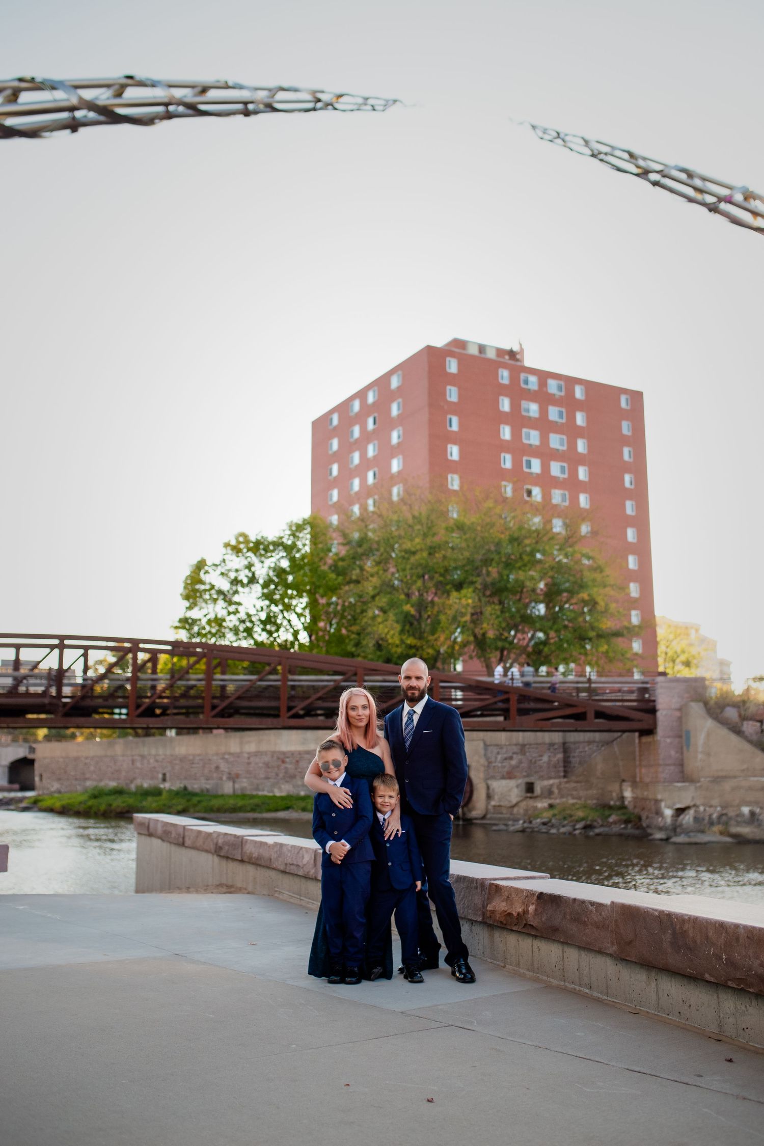 A family in formal wear stands on a riverside path with a brick apartment building and bridge in the background.