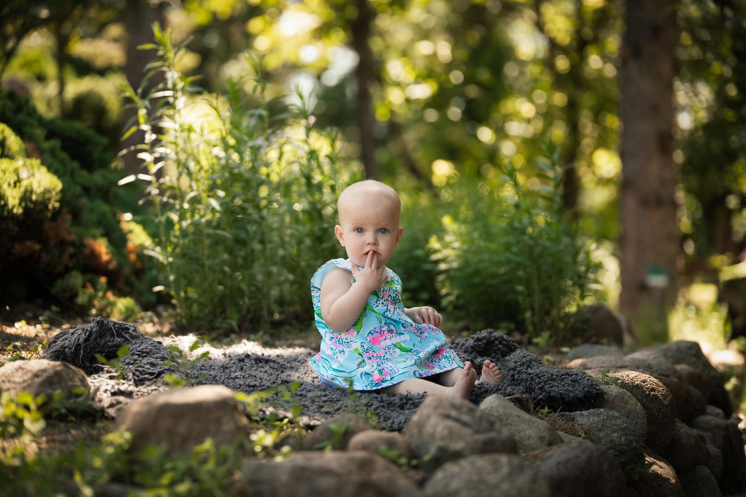 Baby sits on decorative garden rocks amid lush summer greenery and trees.