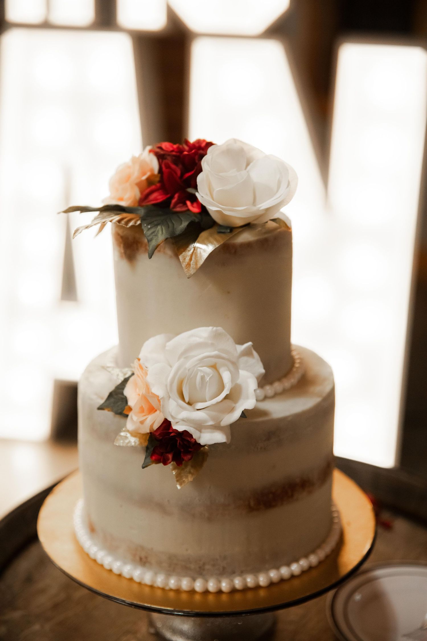 Two-tiered white wedding cake adorned with red and white roses and pearl trim.