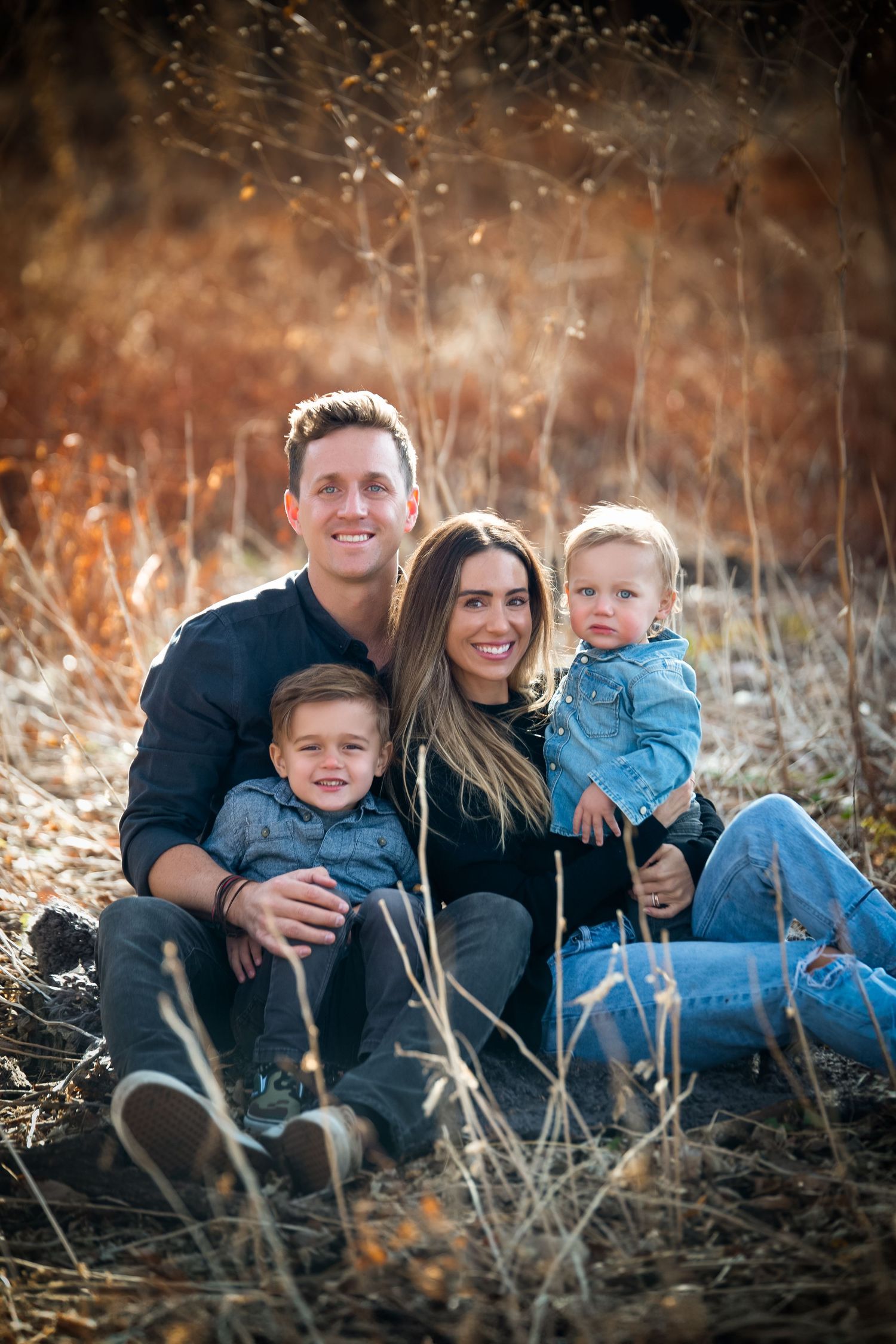 A family in casual attire sits together on autumn grass during a warm outdoor photo session at sunset.