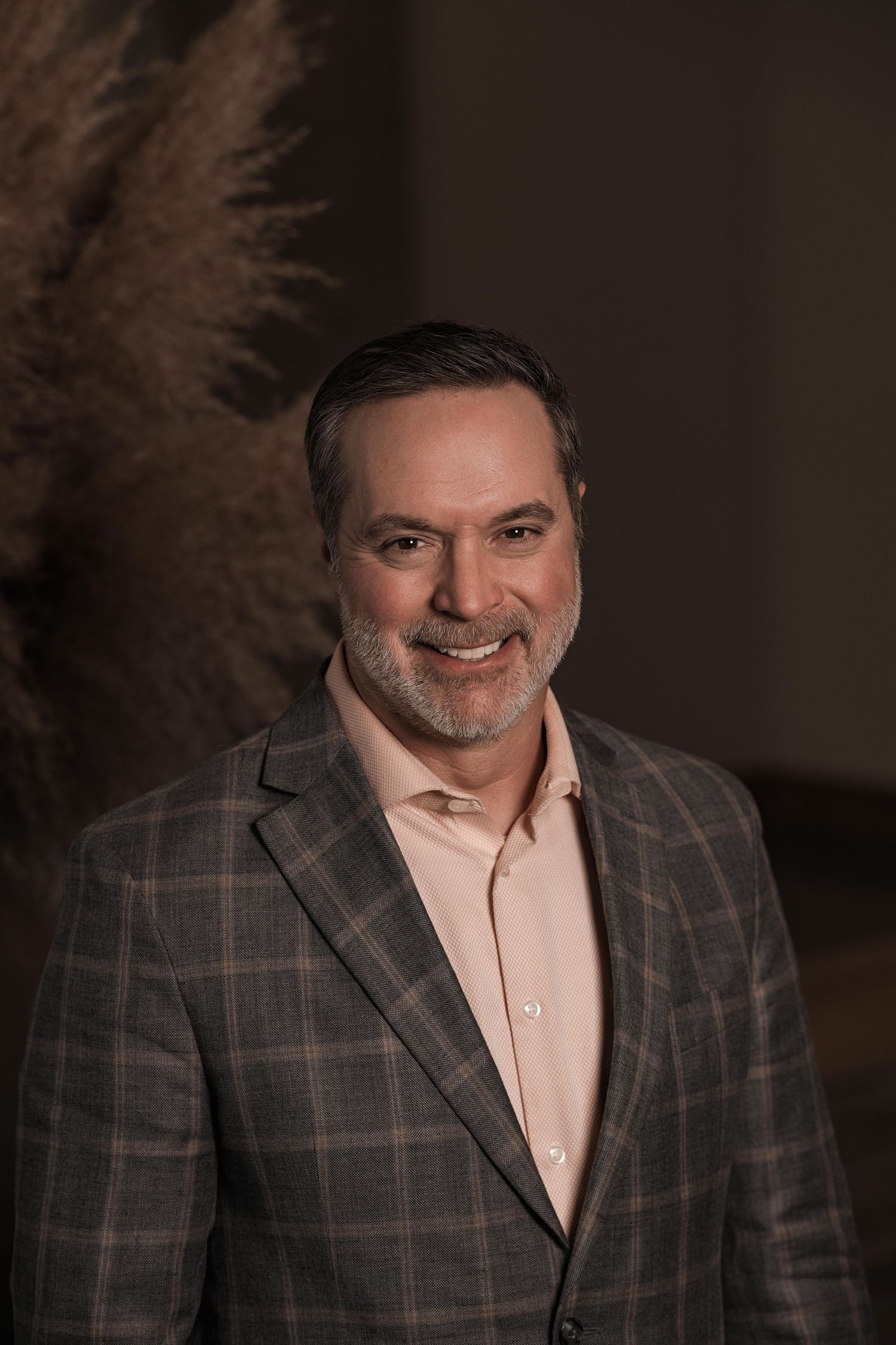 Professional portrait of a person wearing a brown plaid blazer and light colored shirt against a dark background.