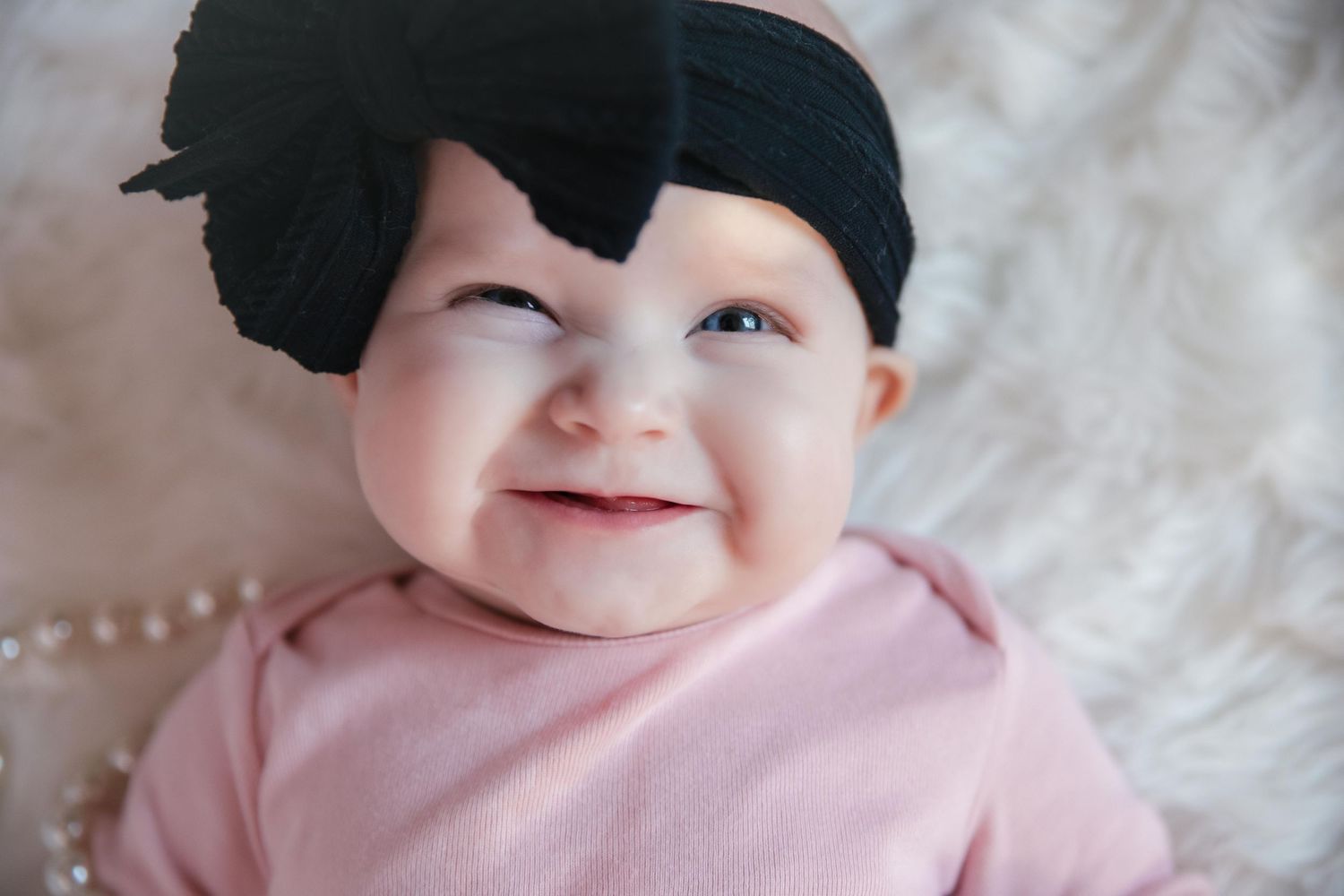 Infant wearing pink outfit smiles while lying on white blanket in heartwarming portrait.