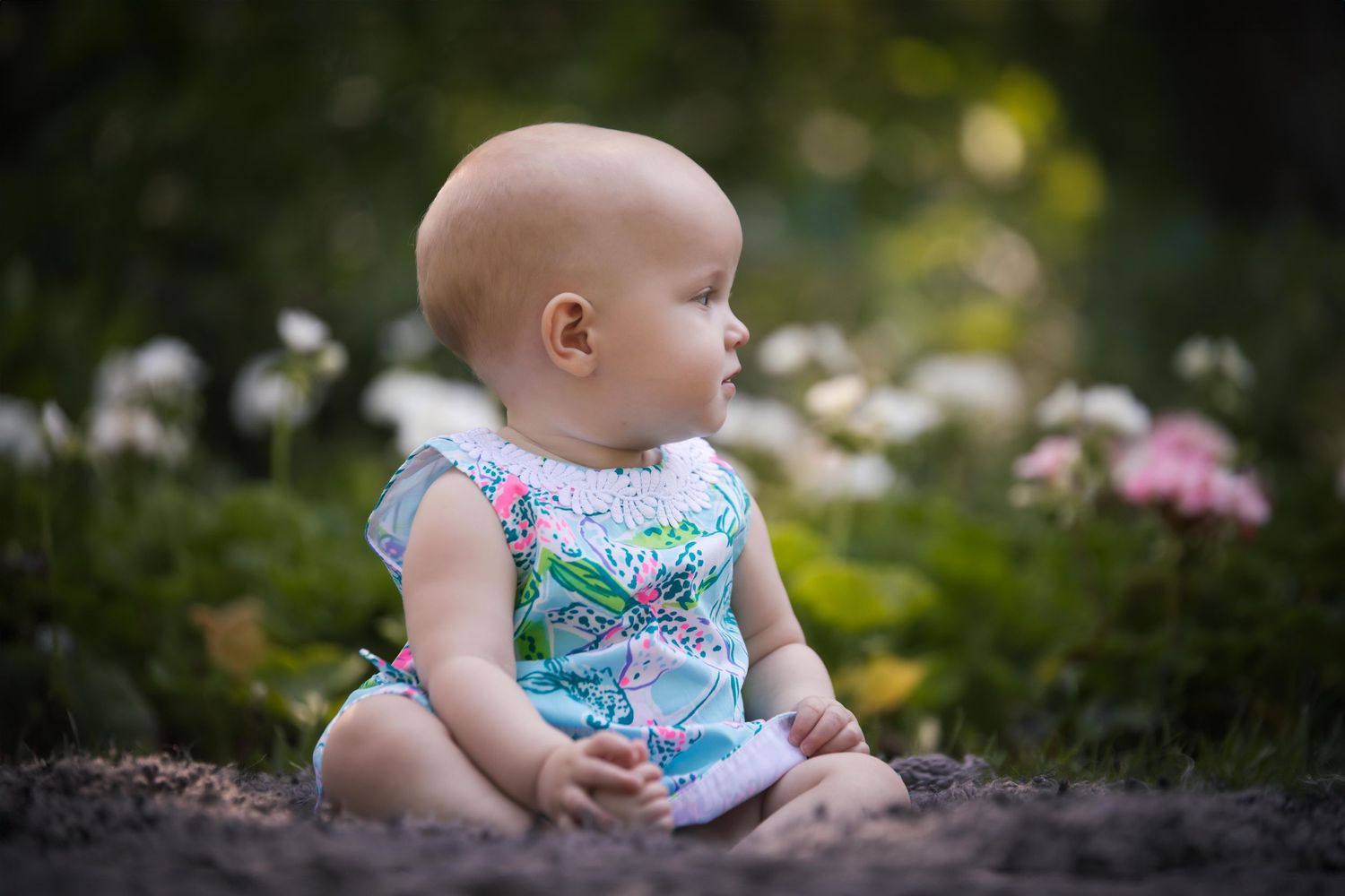 Baby in blue floral dress sits on natural stone landscaping surrounded by greenery.