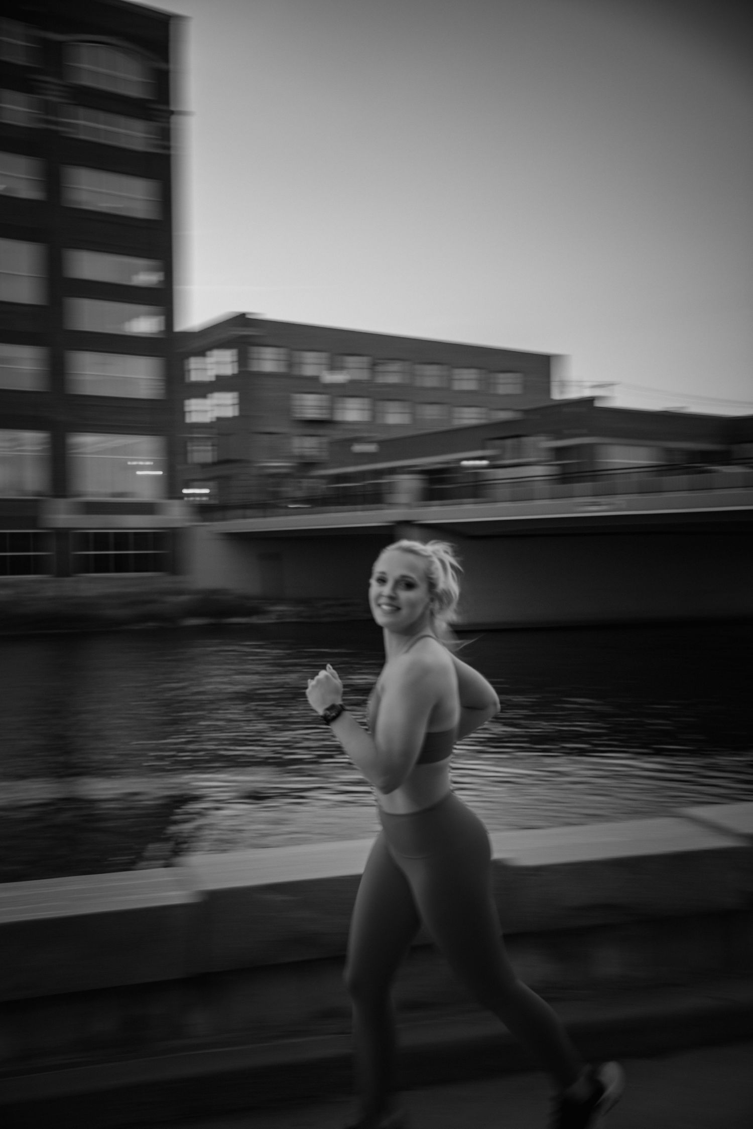 A black and white photo captures someone jogging along an urban waterfront with modern buildings in the background.