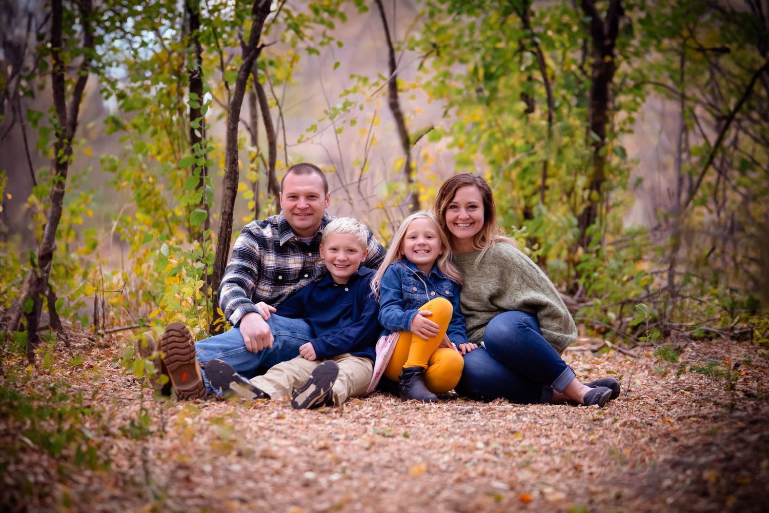 A family sits together on fallen leaves during an autumn photoshoot in a wooded area.