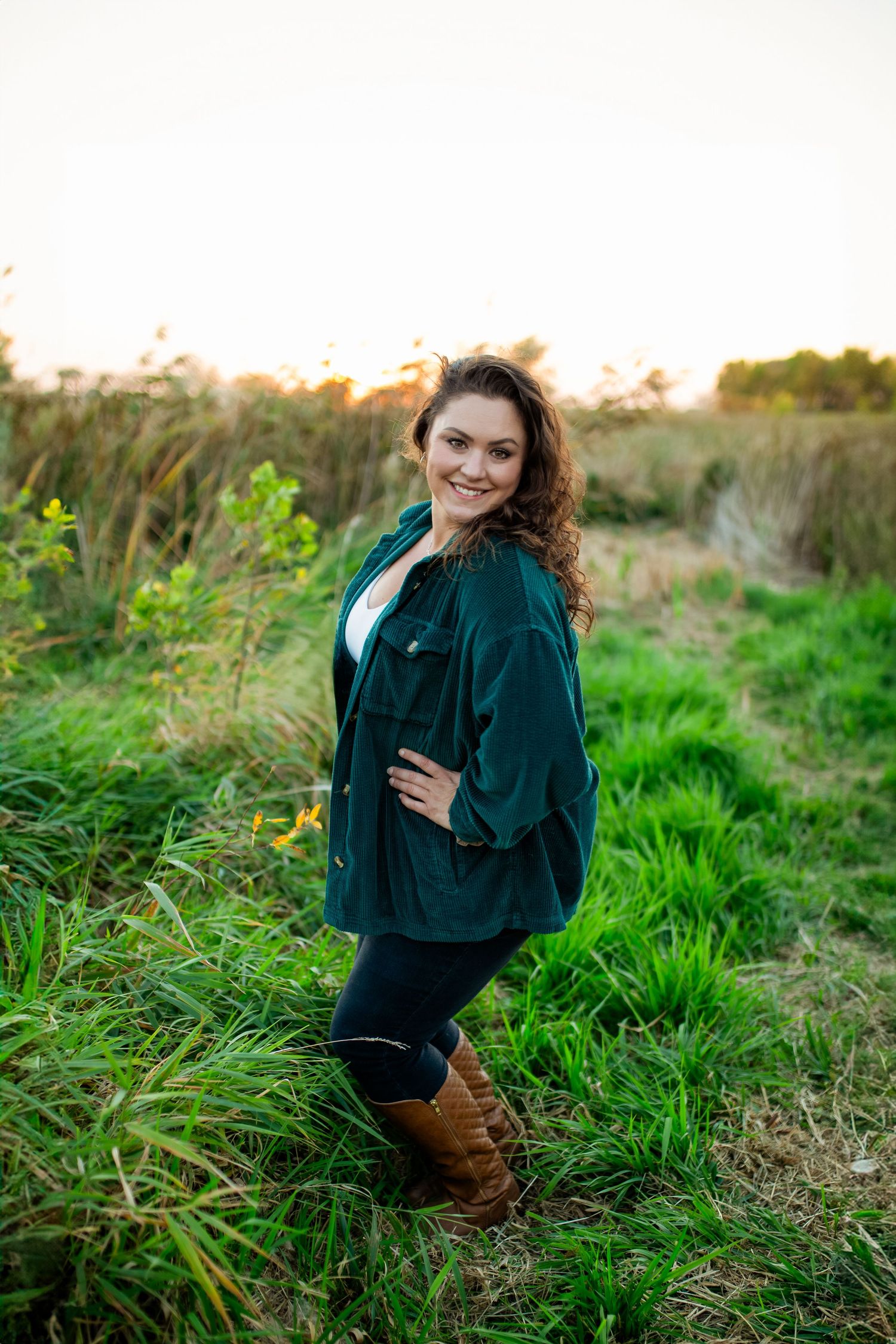 Person wearing dark green sweater sits in tall grass at sunset in outdoor portrait session.