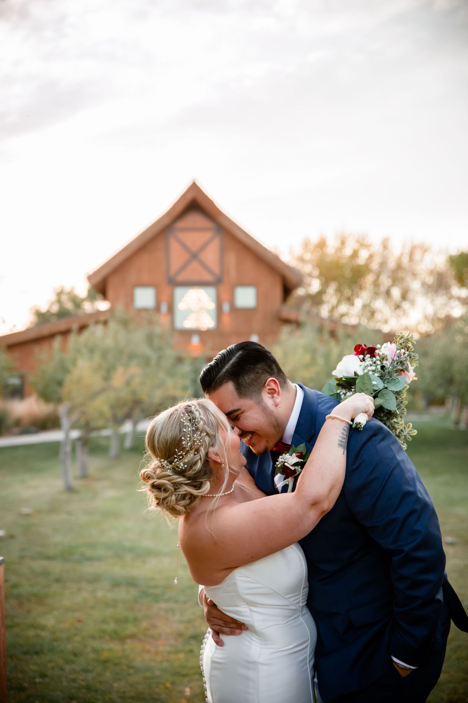 Romantic wedding couple shares intimate kisses in front of rustic barn buildings during golden hour.