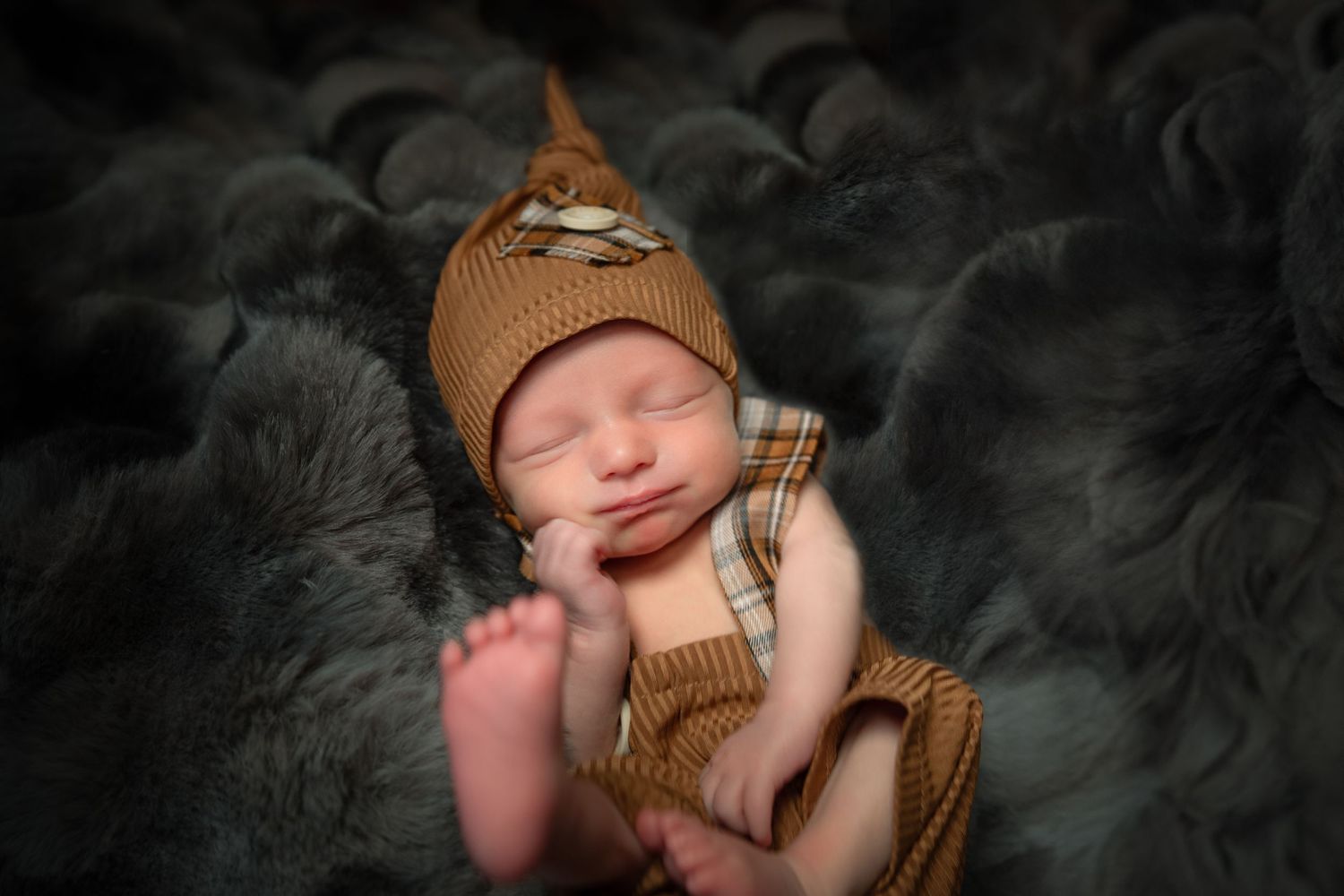 Sleeping newborn wearing brown knit hat and plaid overalls rests peacefully on soft gray blanket.