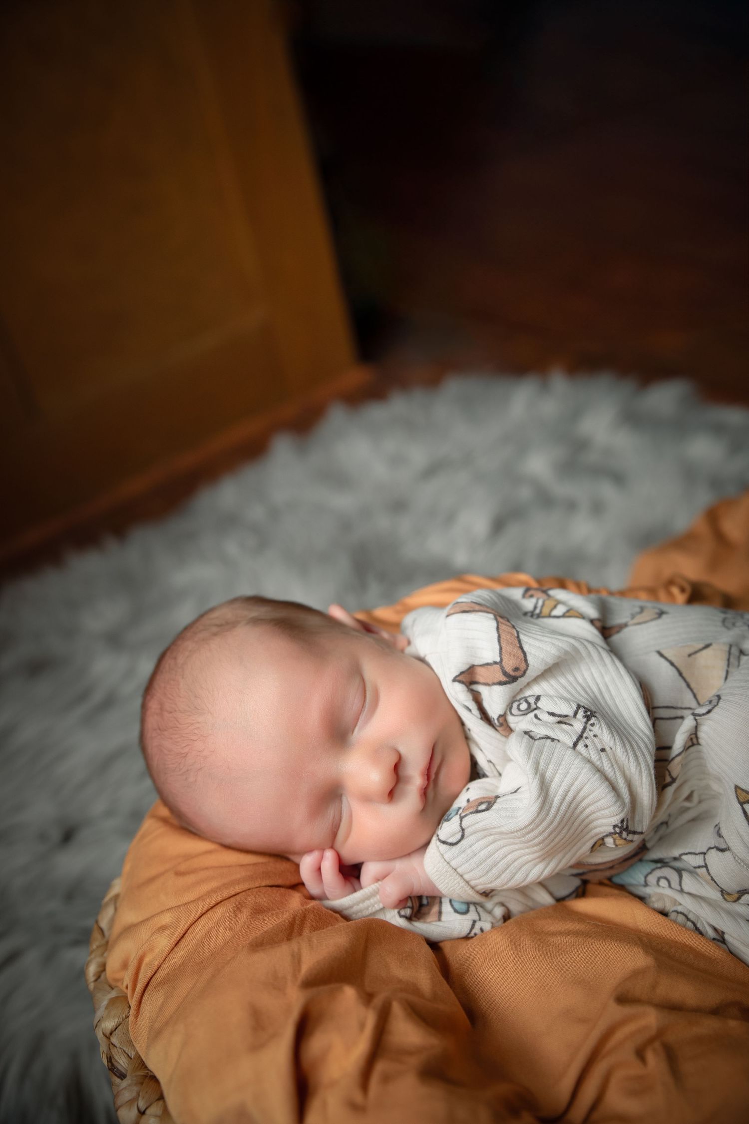 A sleeping baby wearing a white patterned onesie rests peacefully on brown fabric bedding.