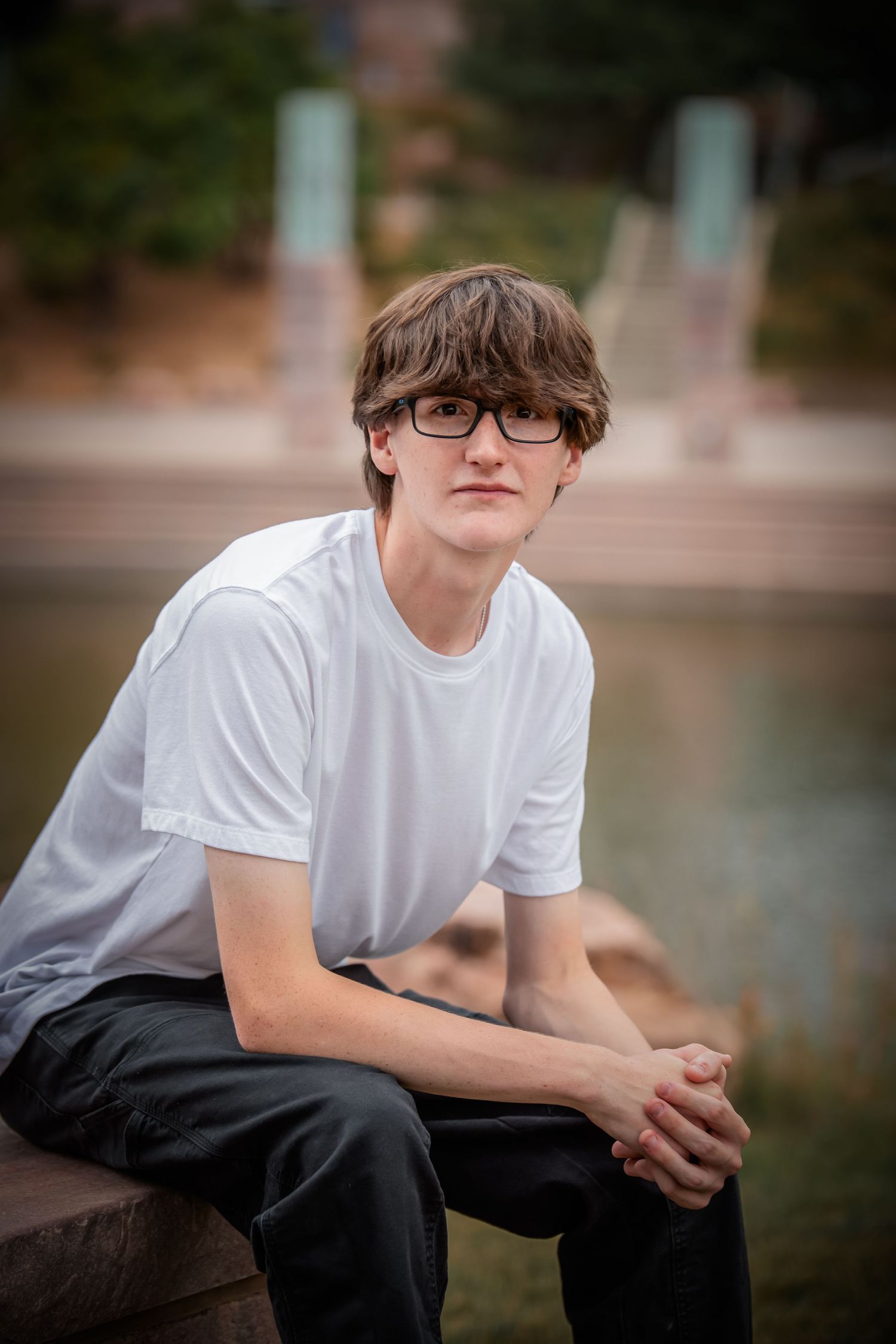 Youth wearing glasses and a white t-shirt sits thoughtfully on an outdoor bench near water.