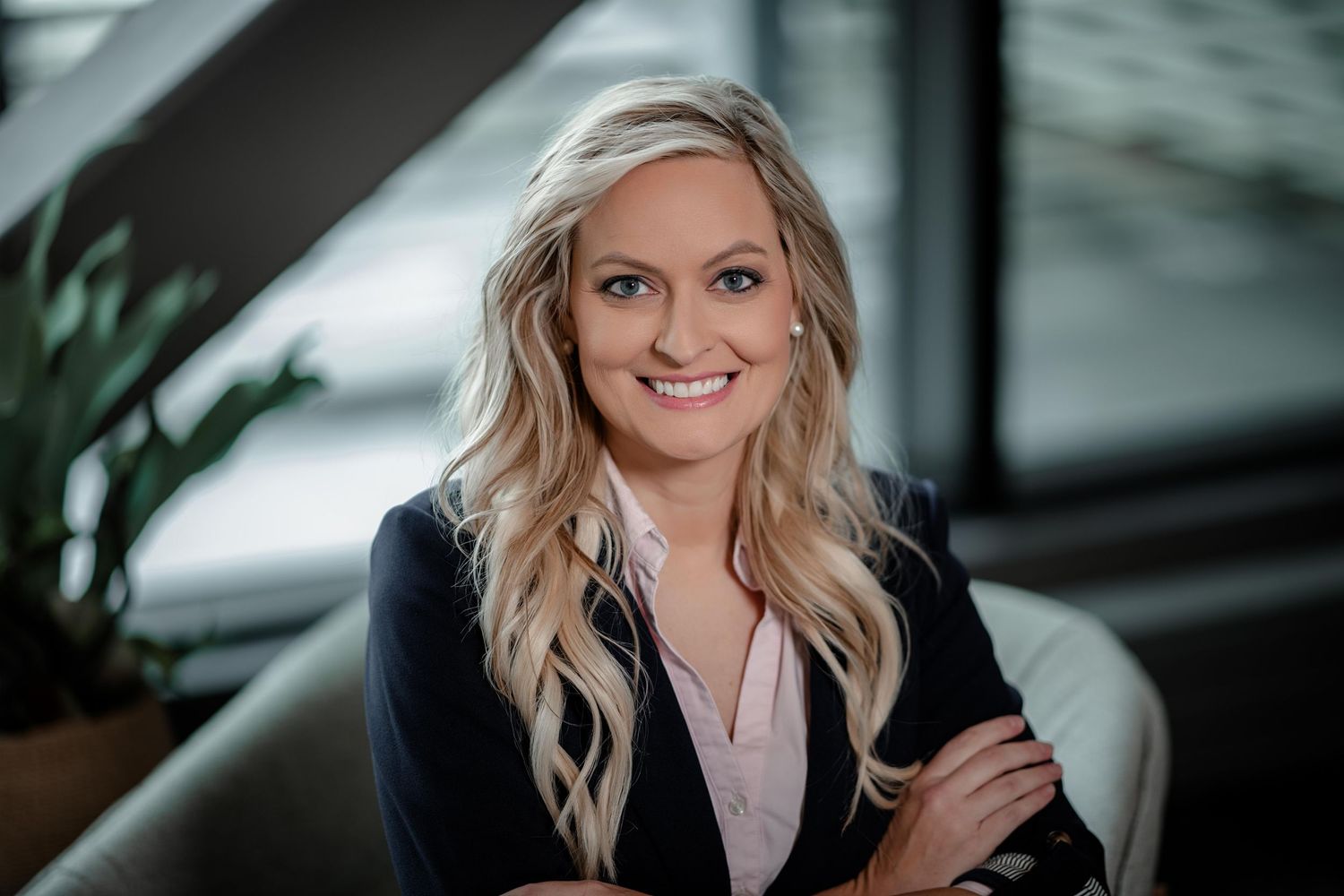 Professional headshot of a person with blonde wavy hair wearing a navy blazer and light blouse, smiling warmly at camera.