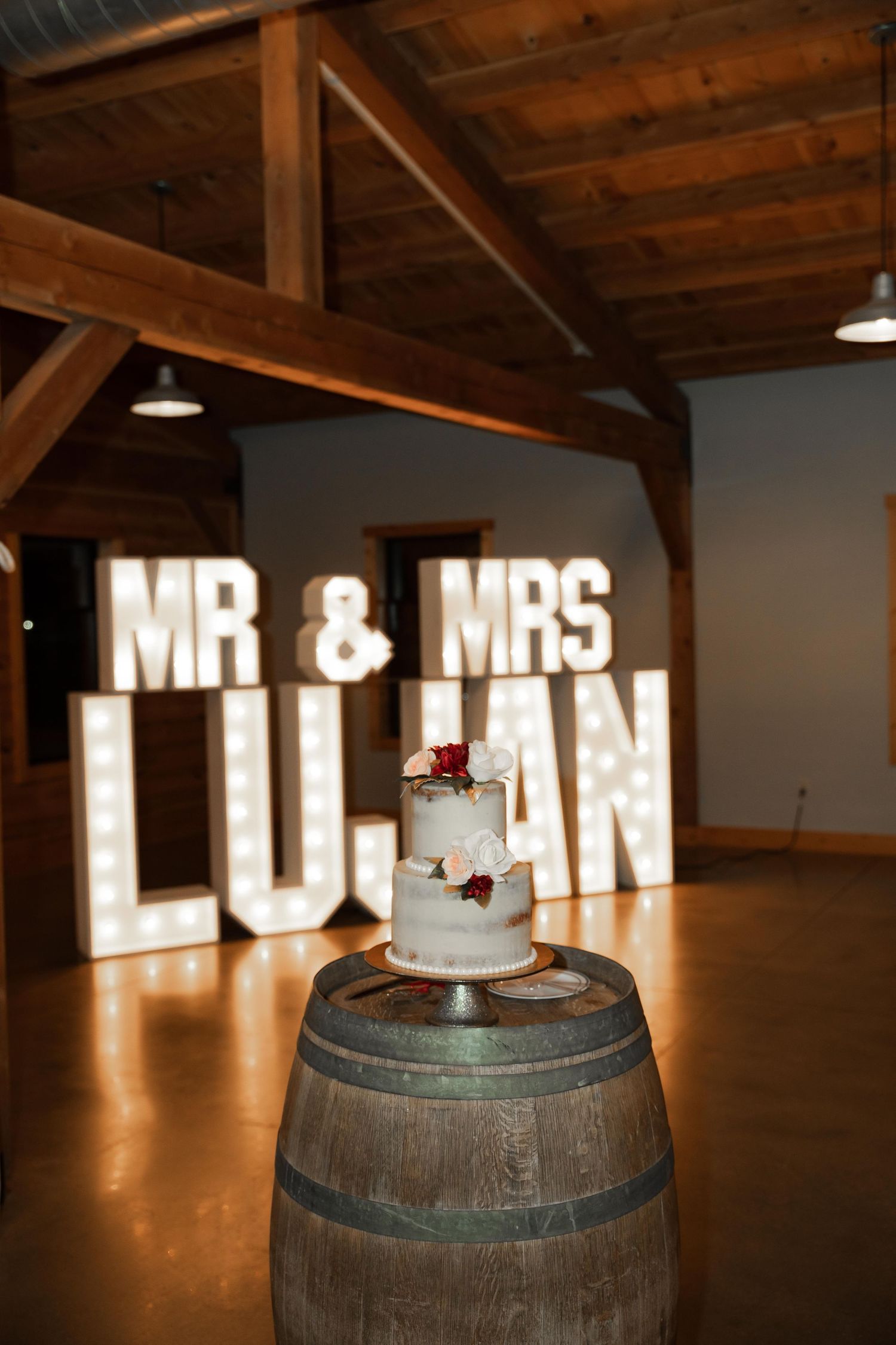 Wedding cake sits atop wooden barrel with glowing marquee letters in rustic indoor venue. Photographer in Sioux Falls, SD