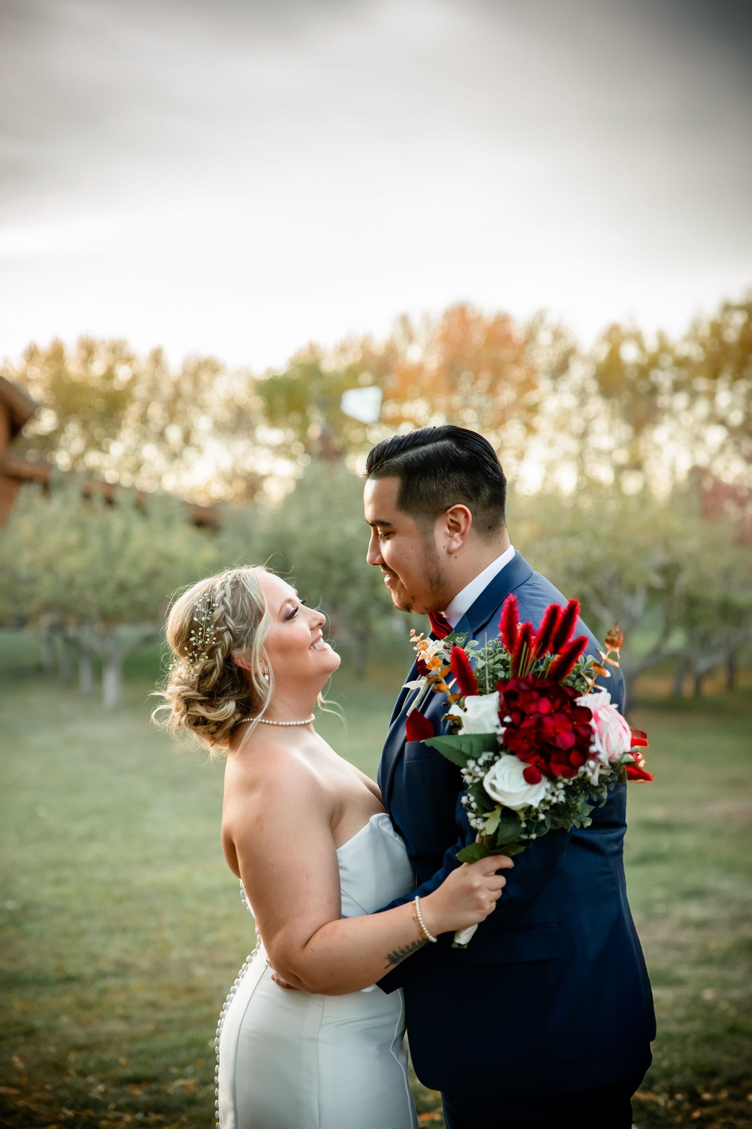 A romantic wedding couple shares tender moments in a sunset field with red and white bouquet.