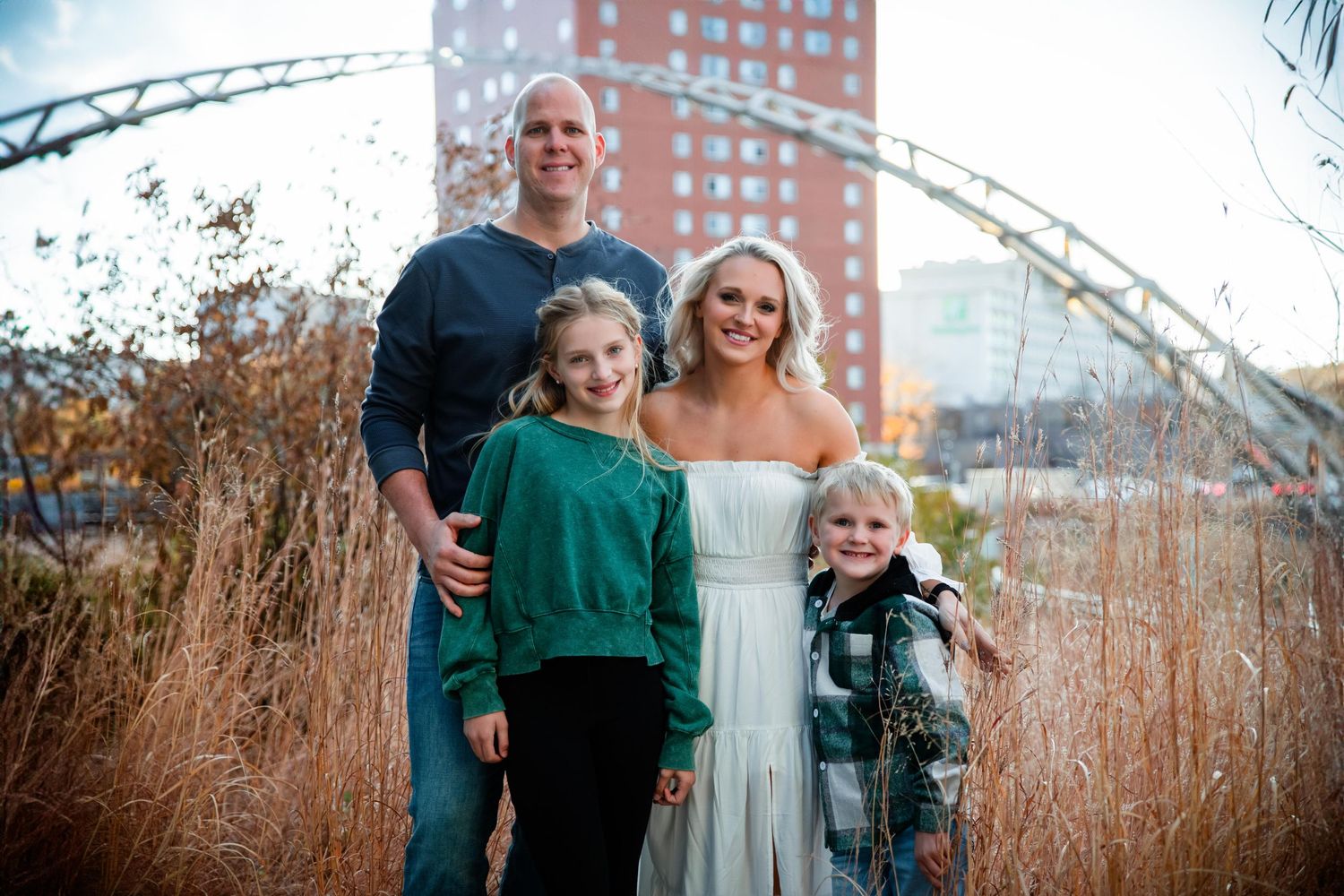 A group stands together in front of tall grass with a brick building and metal arch visible behind them.