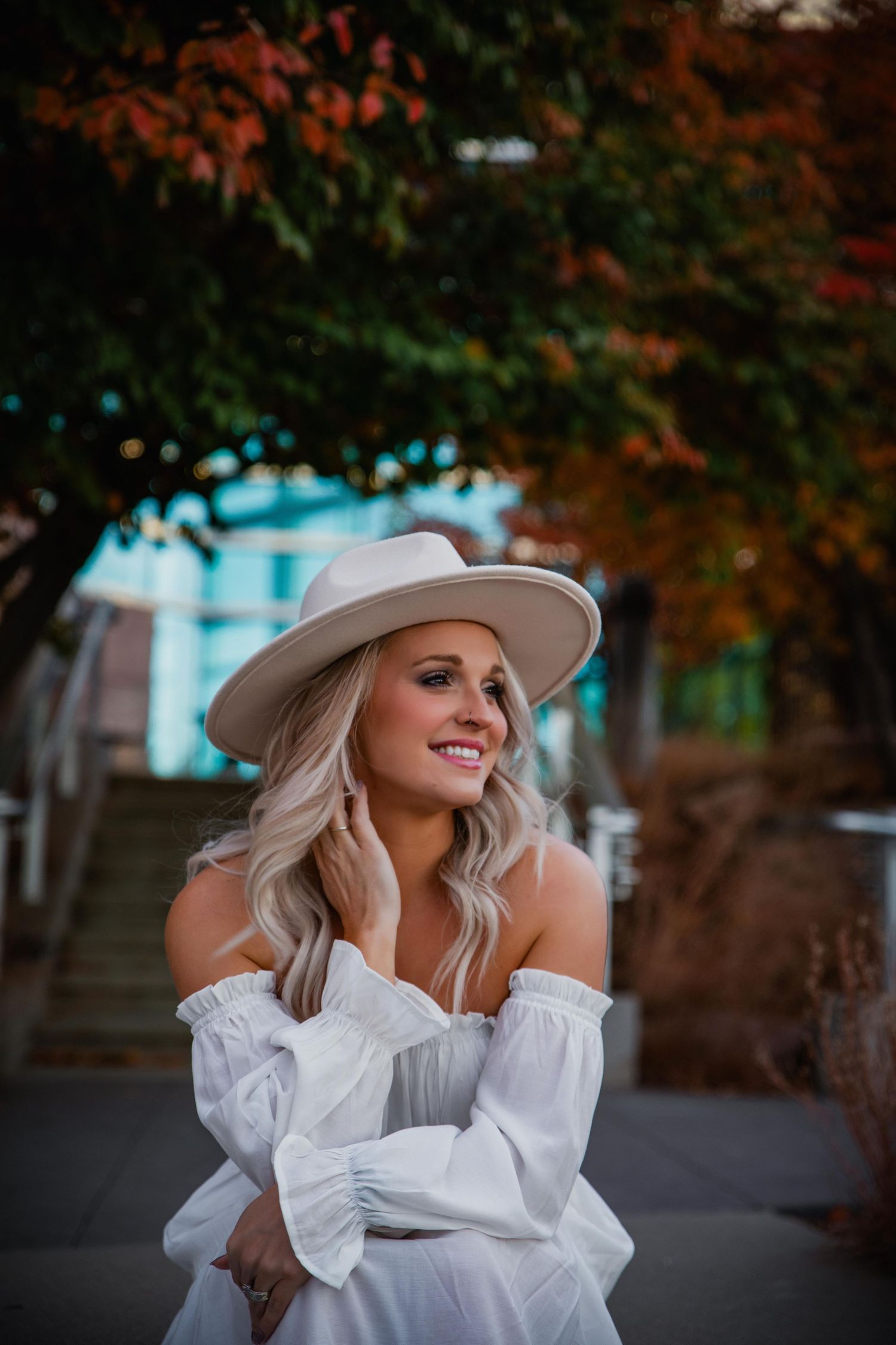 A woman in a white blouse and beige hat poses outdoors during autumn with orange foliage in the background.