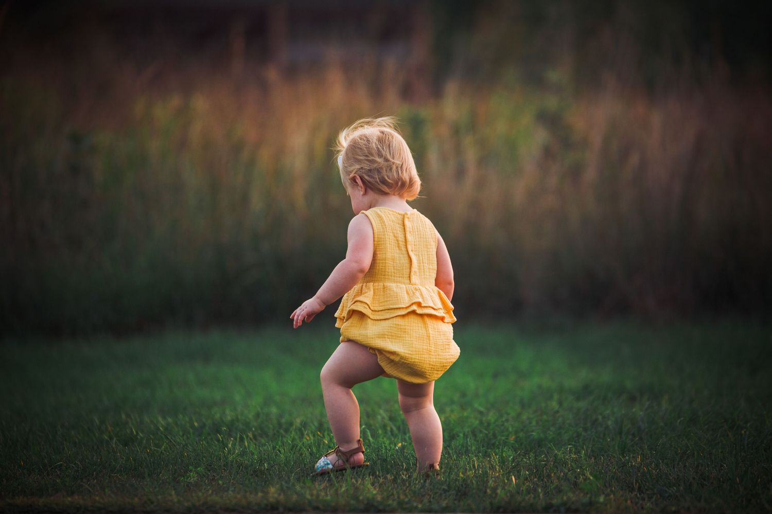 A small figure in a yellow romper walks on grass during golden hour with tall grass in the background.
