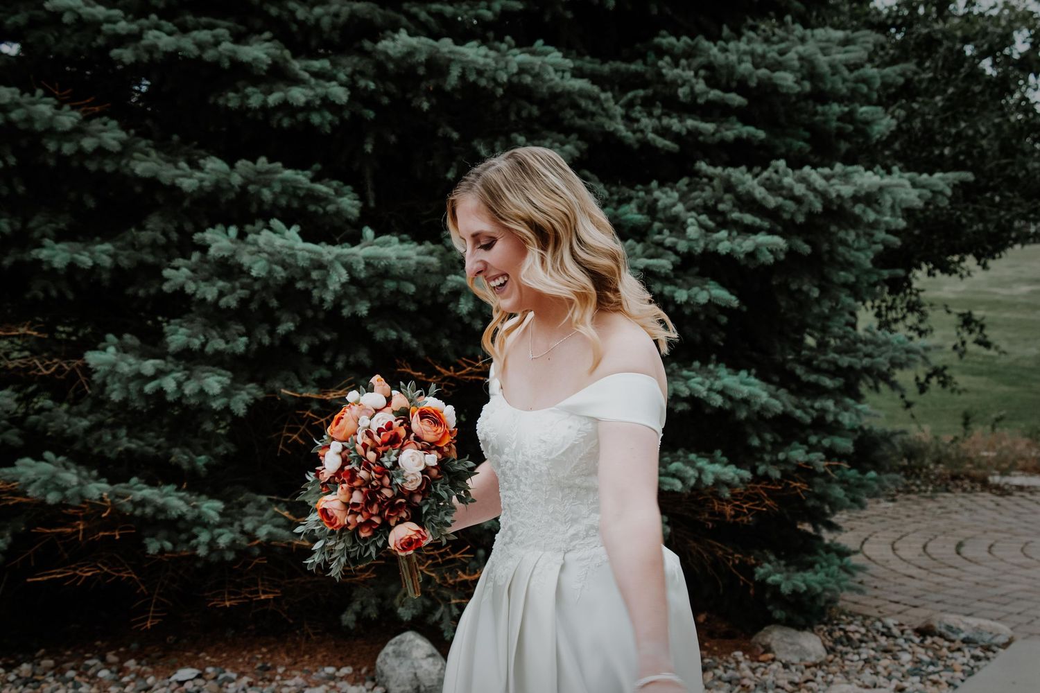 A bride in an elegant off-shoulder gown holds a vibrant autumn bouquet against evergreen trees.