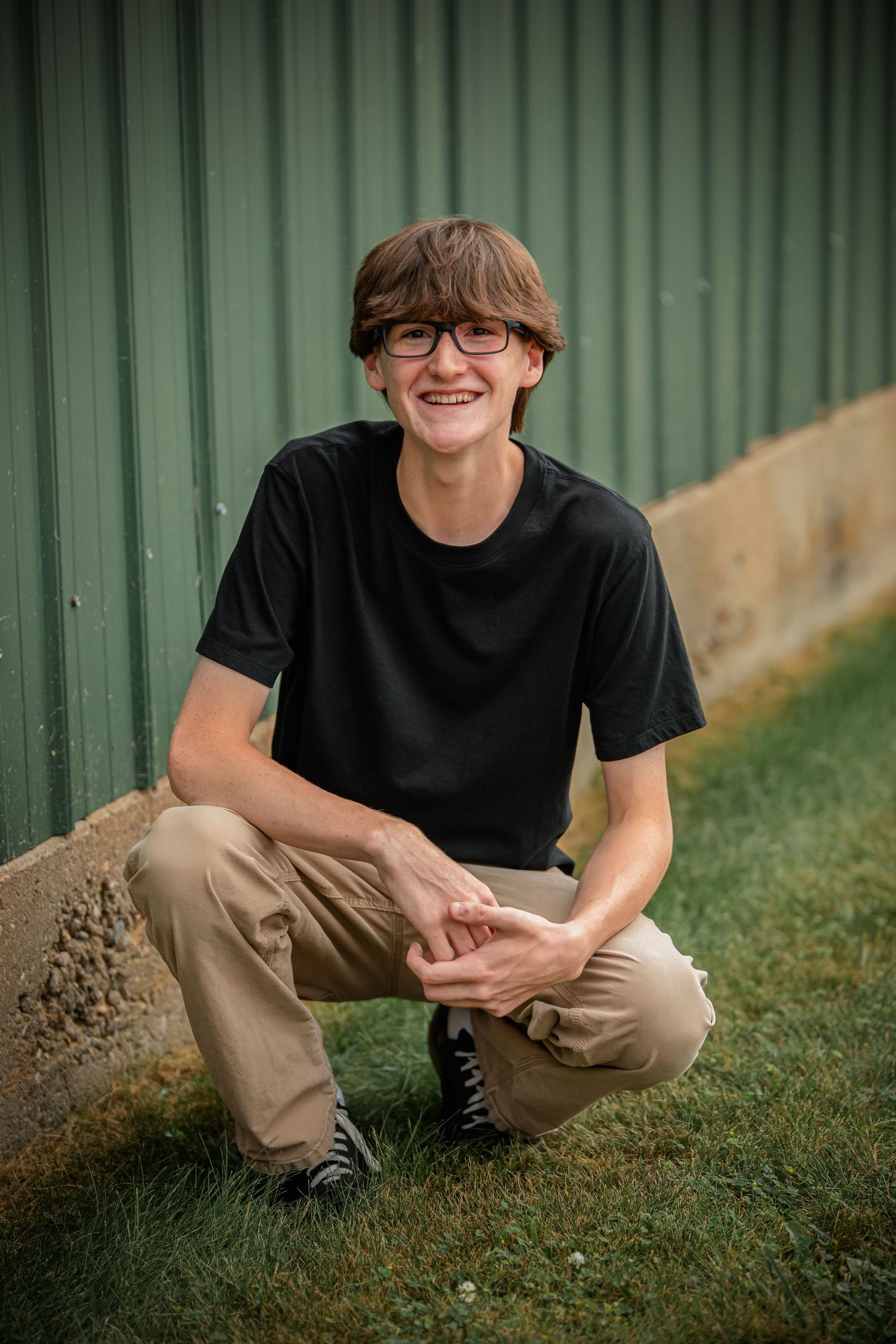 Person in black shirt and khaki pants sitting on grass with green corrugated wall background.