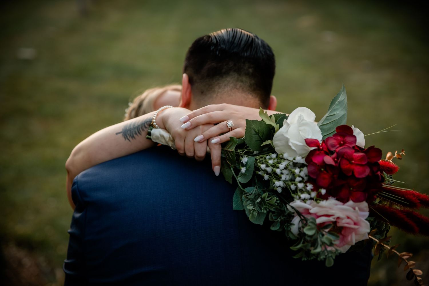 A tender embrace between two people at an outdoor wedding, with a beautiful bouquet of red and pink roses visible.