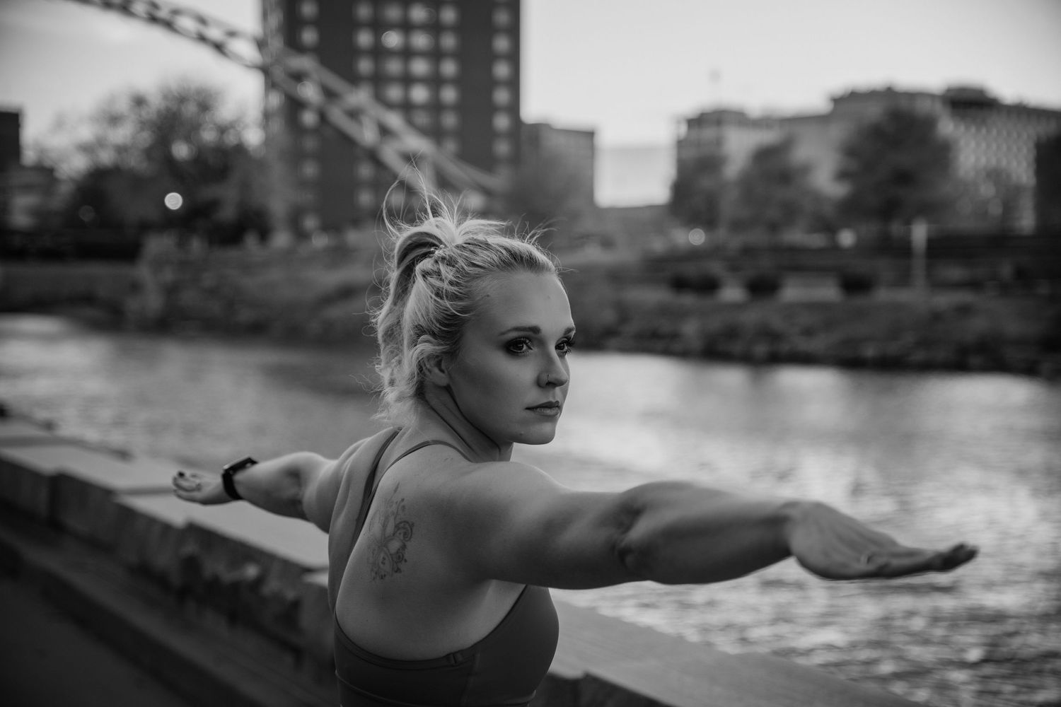 A black and white photo of a dancer stretching with arms outstretched by a waterfront with urban buildings in background.