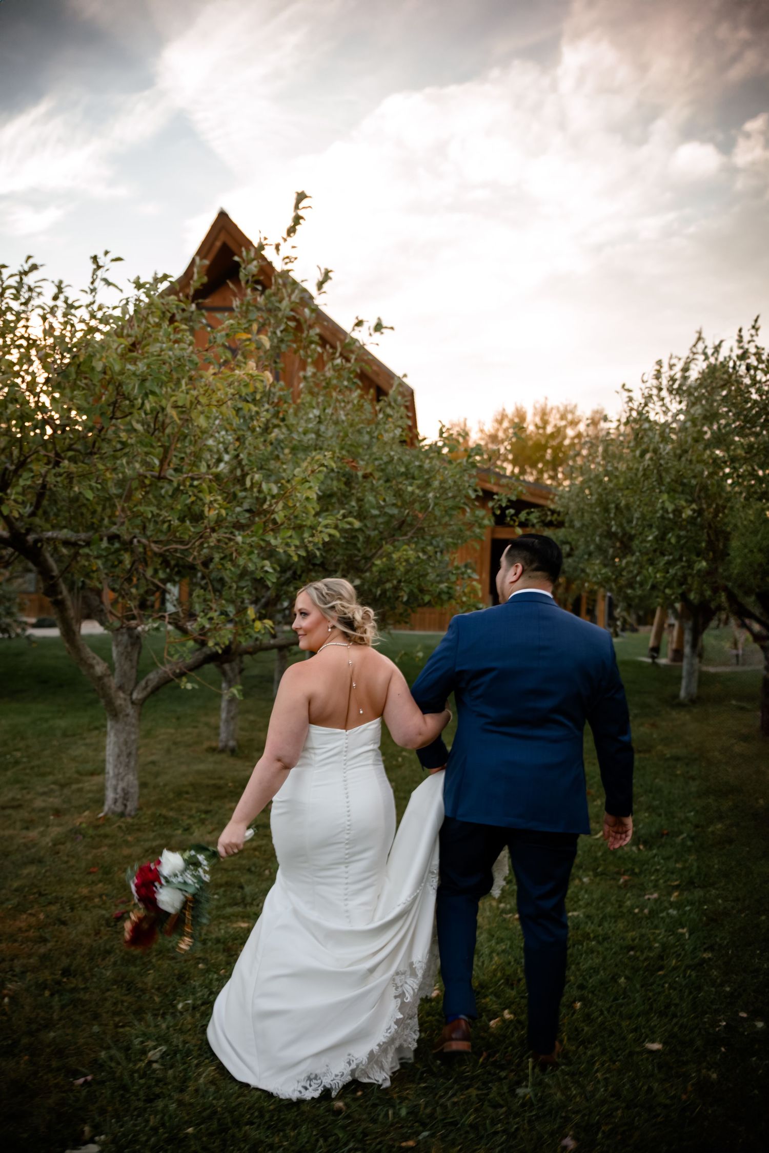 A couple in wedding attire walks through an apple orchard at sunset during their romantic autumn celebration.