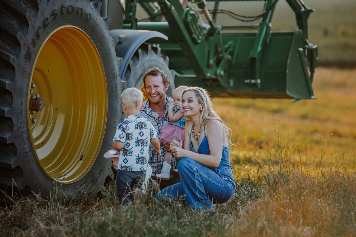 Family enjoying a rustic photo shoot in a golden field at sunset with tractors in the background.