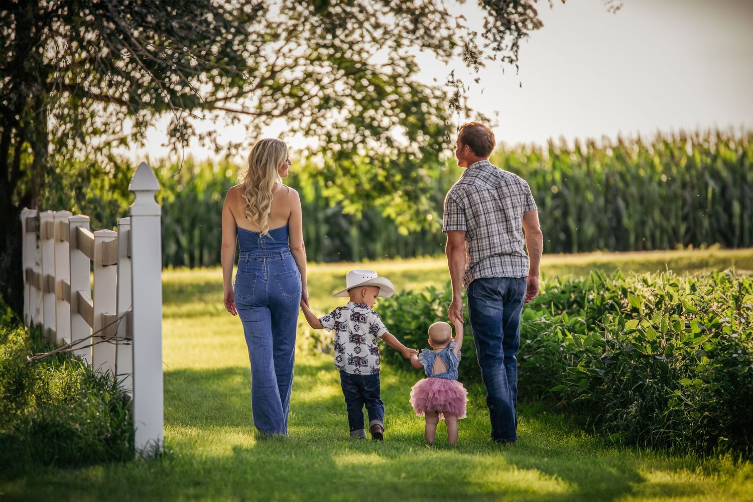 Family walking together along white fence near cornfield during golden hour.