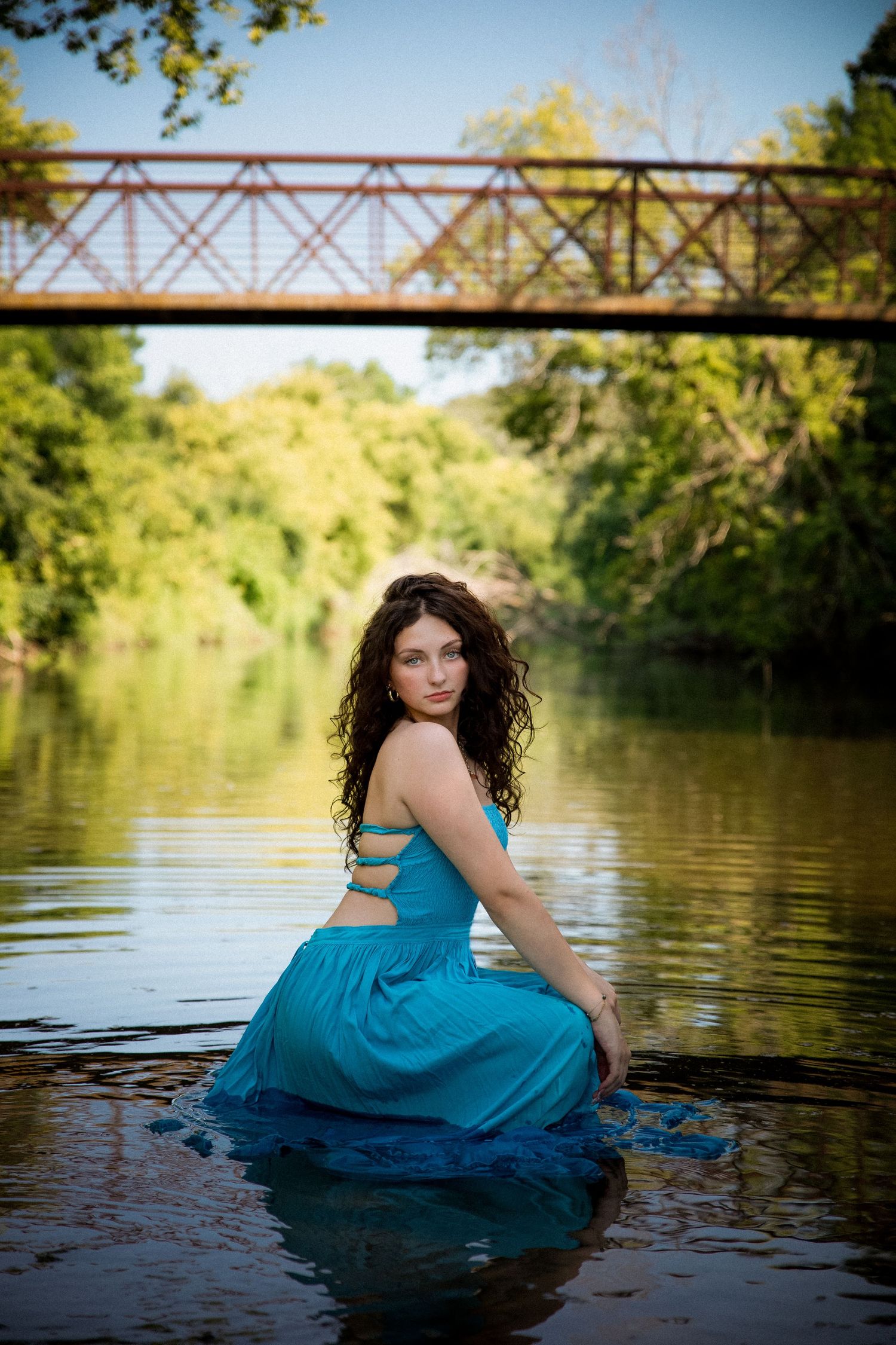 Young woman in teal dress sitting in a river with a rustic iron bridge and lush green trees in the background.