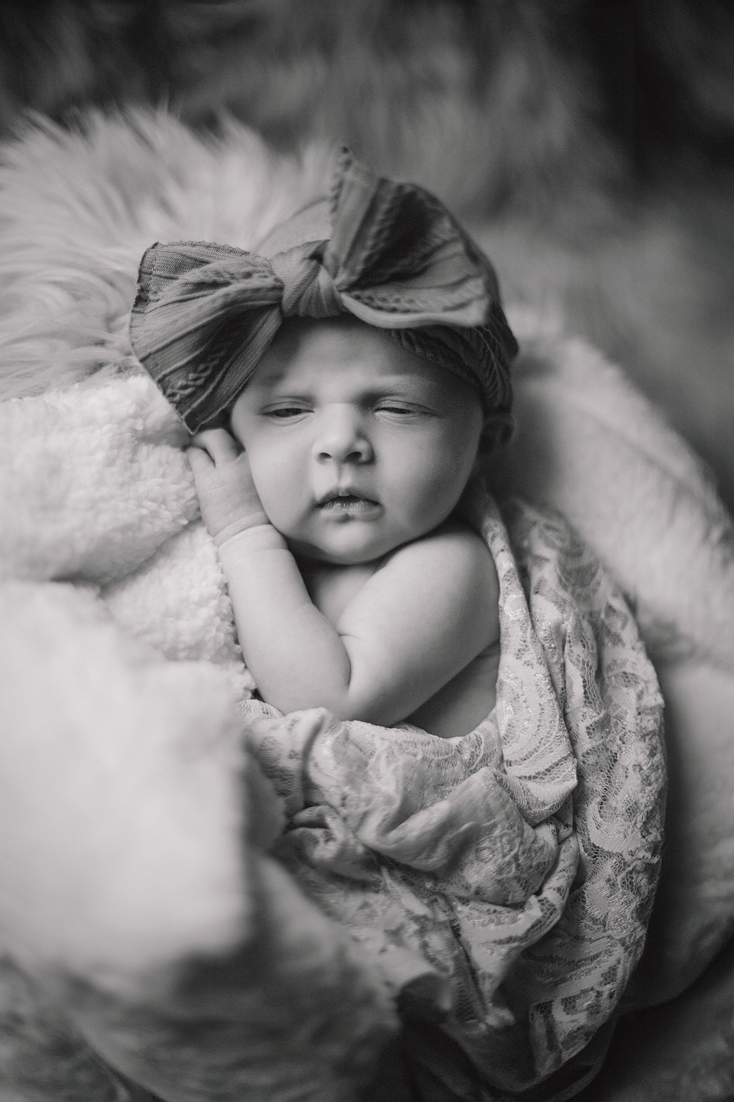 Black and white portrait of baby wearing lace dress and bow on soft fur backdrop.