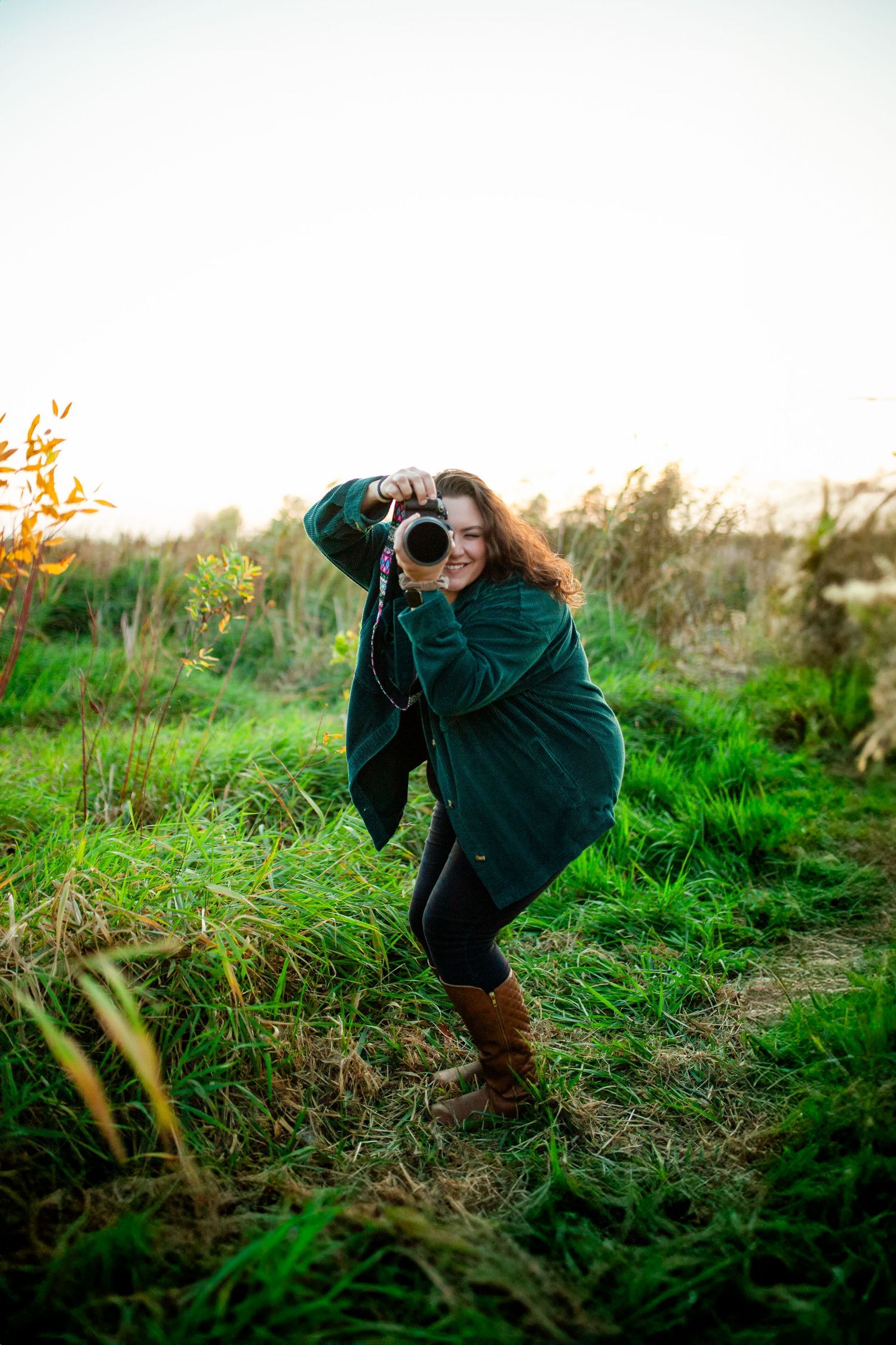 A photographer in a dark jacket crouches low to get a special angle while taking pictures in a grassy field at sunset.