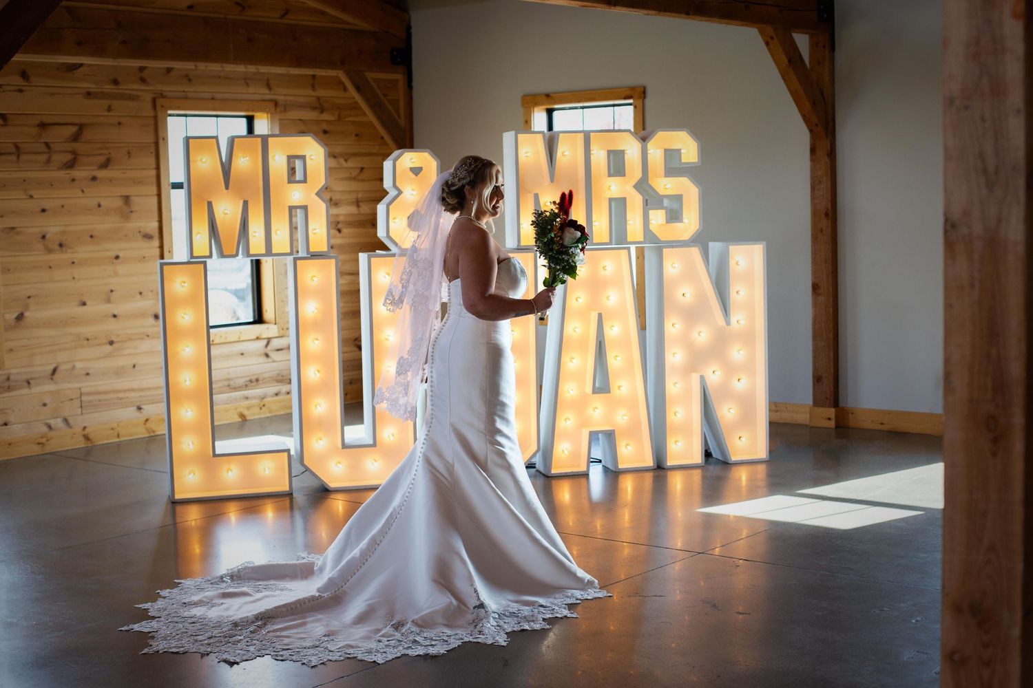 Wedding dress silhouette against illuminated 'MR & MRS' marquee letters in rustic wooden venue.