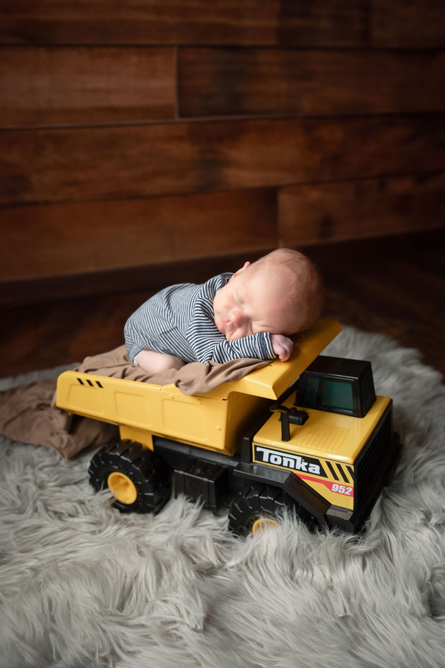 A baby lies on a fluffy white rug next to yellow toy construction vehicles in a rustic wood-paneled room.