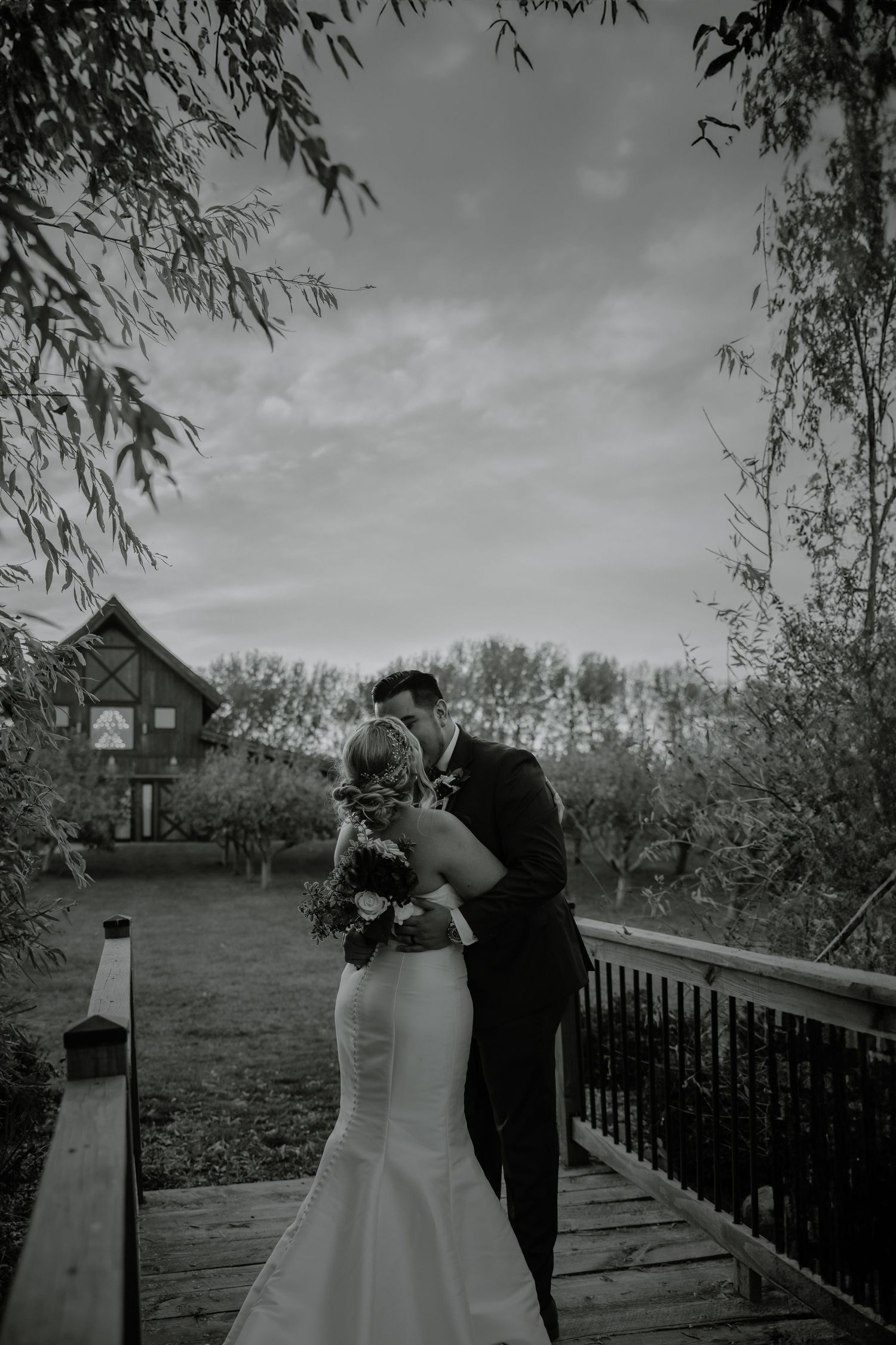 Black and white wedding photo of couple embracing on wooden bridge with rustic venue backdrop. Photographer in Sioux Falls, SD