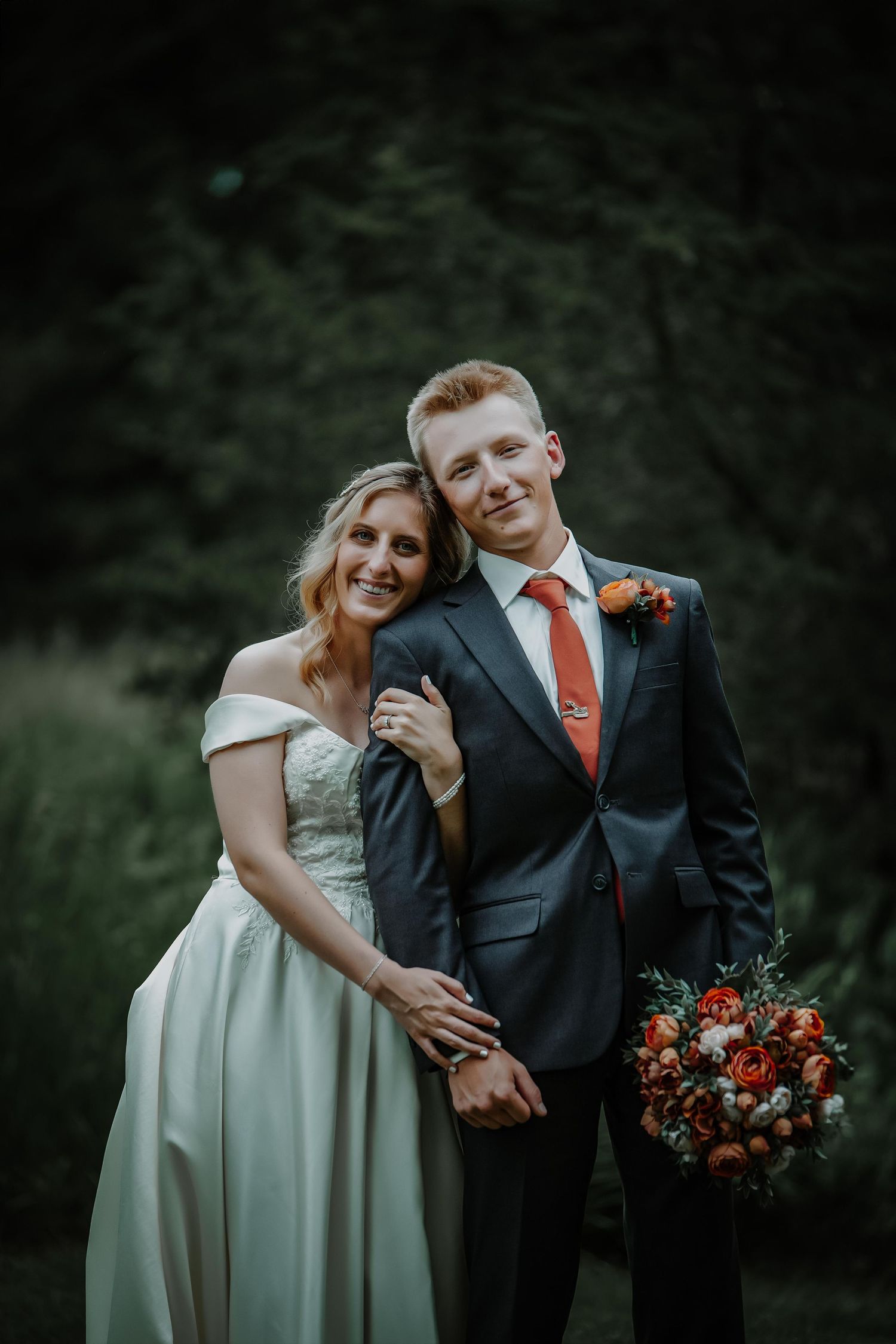 Wedding couple poses together with coral bouquet against moody forest backdrop.