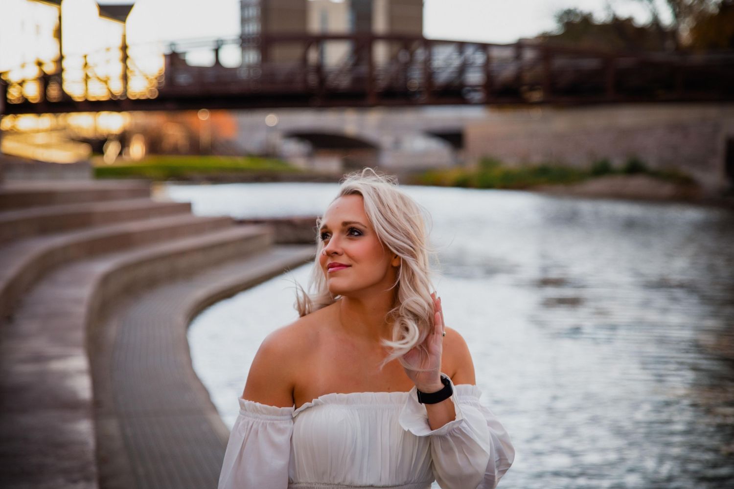 A woman in a white off-shoulder dress poses near a waterfront during golden hour.