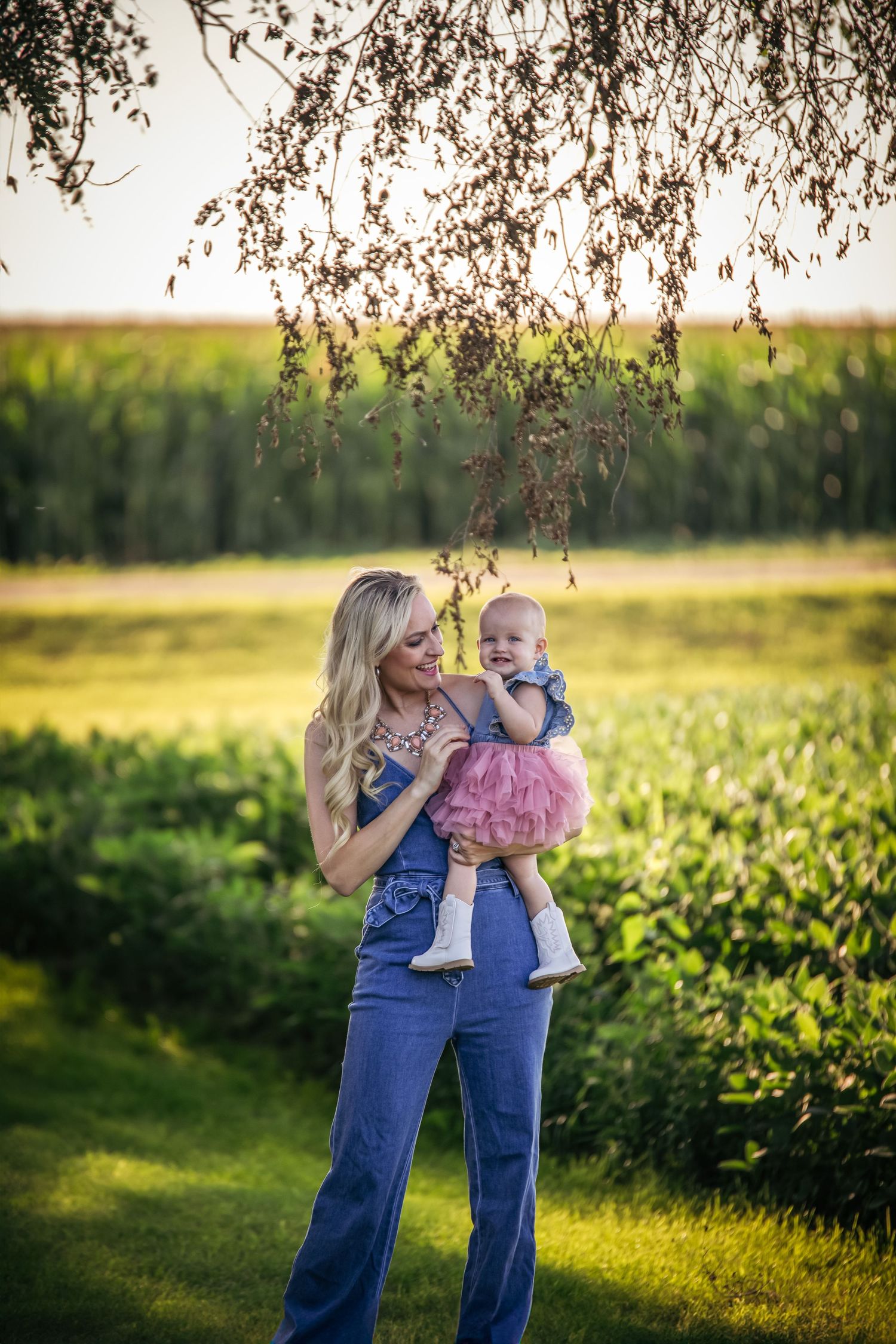 Woman in overalls holds small child against sunset-lit rural backdrop.