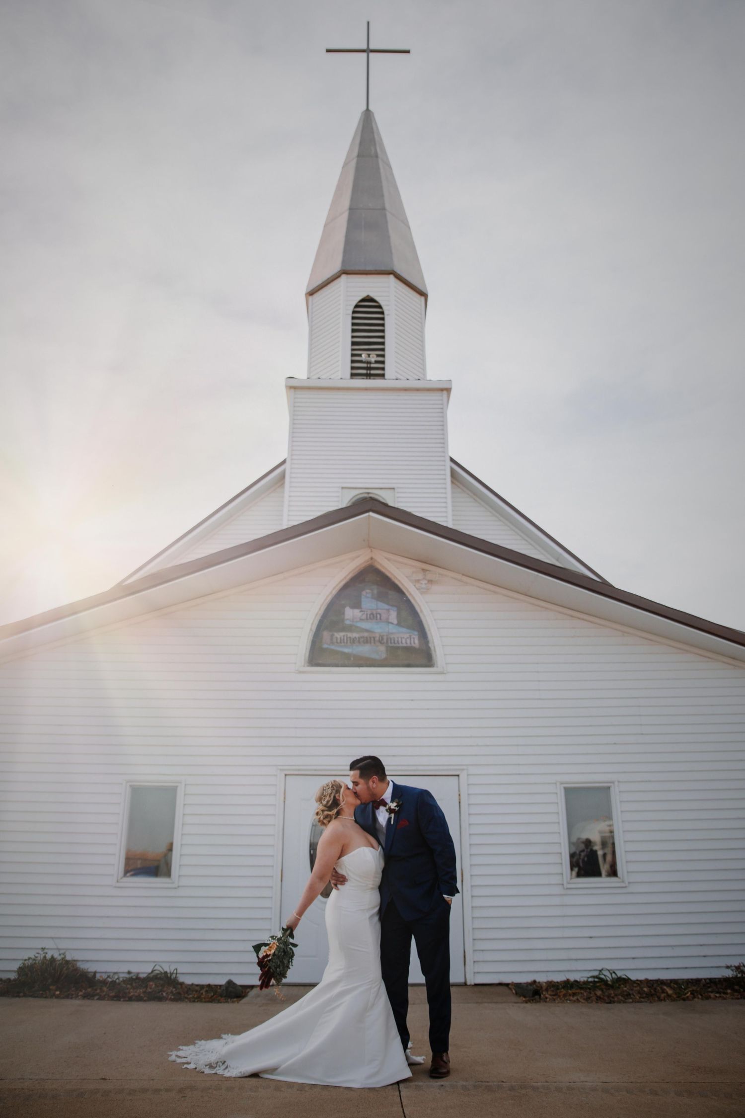 White country church exterior with wedding couple sharing intimate moment against rustic backdrop.