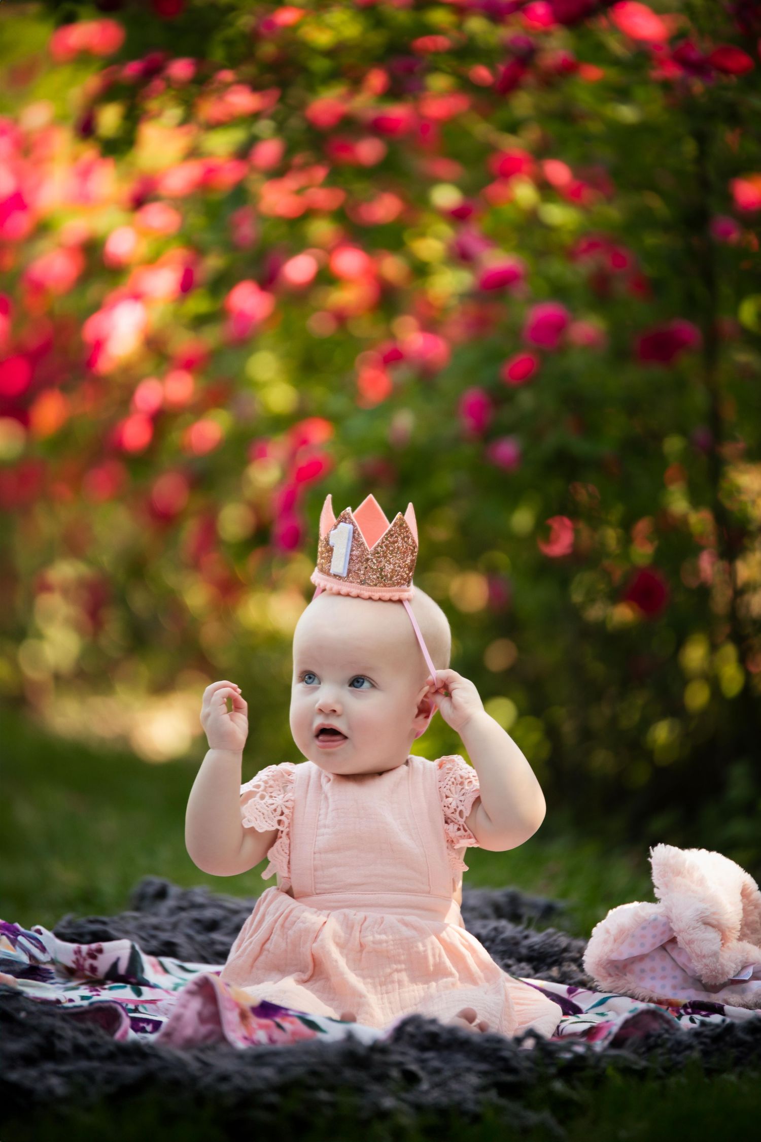 Baby wearing a delicate pink crown sits on a blanket surrounded by vibrant pink flowers in a garden setting.