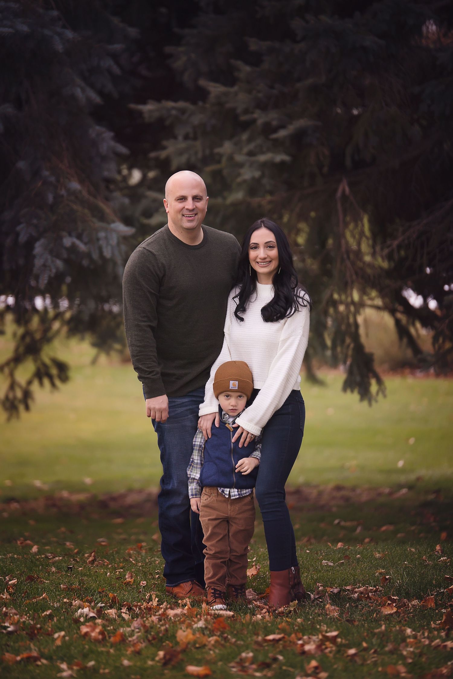 A family of three poses together outdoors in autumn, standing on fallen leaves near evergreen trees.