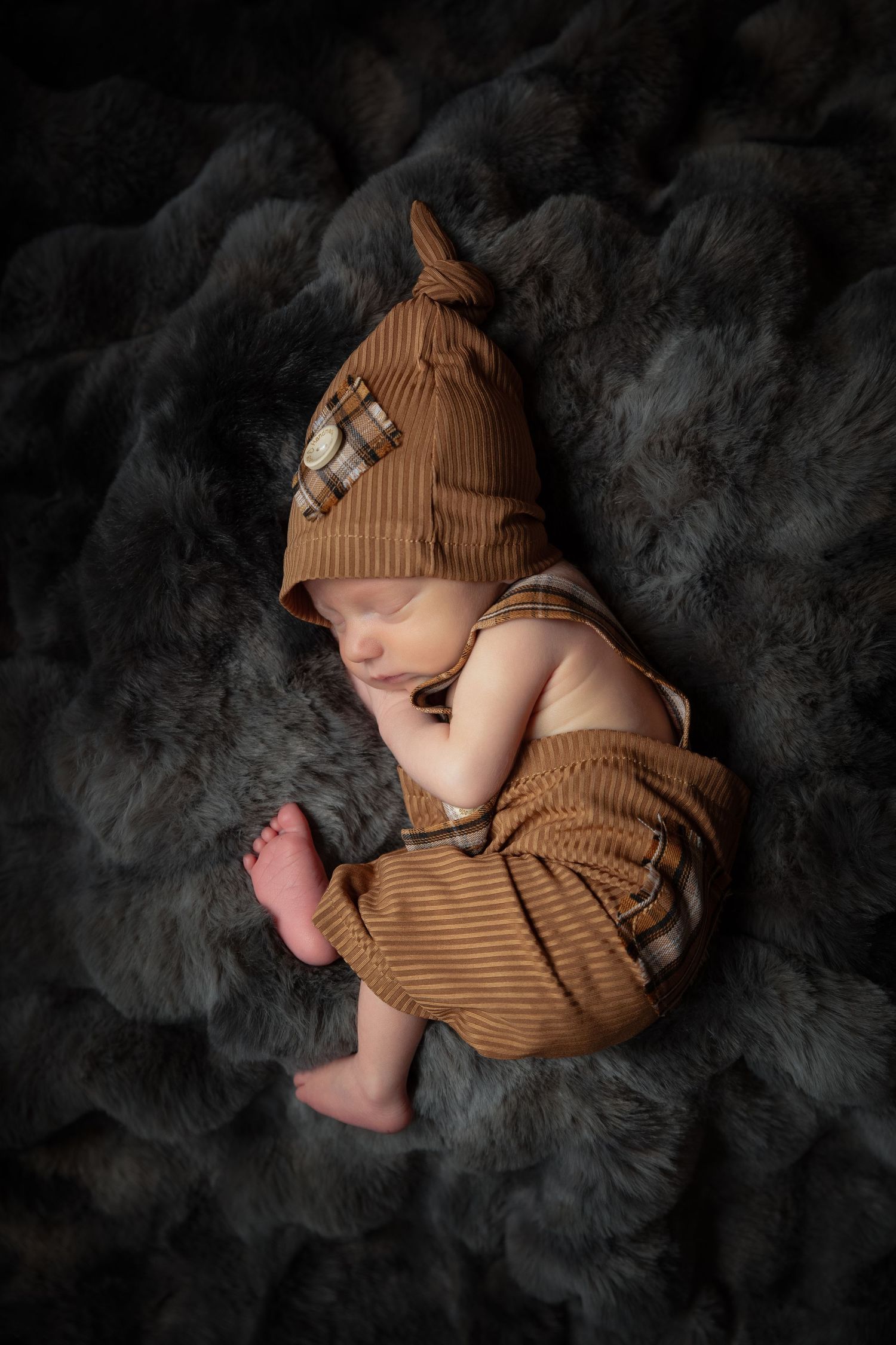 A sleeping baby wearing a brown knit outfit curls up on a dark textured blanket in a series of artistic newborn photos.