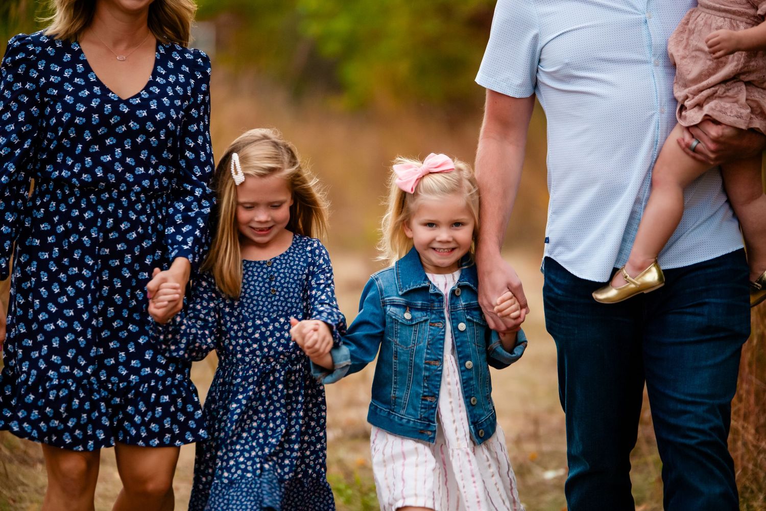 Two young girls holding hands with parents, wearing matching navy polka dot dresses, walking on a wooded trail.