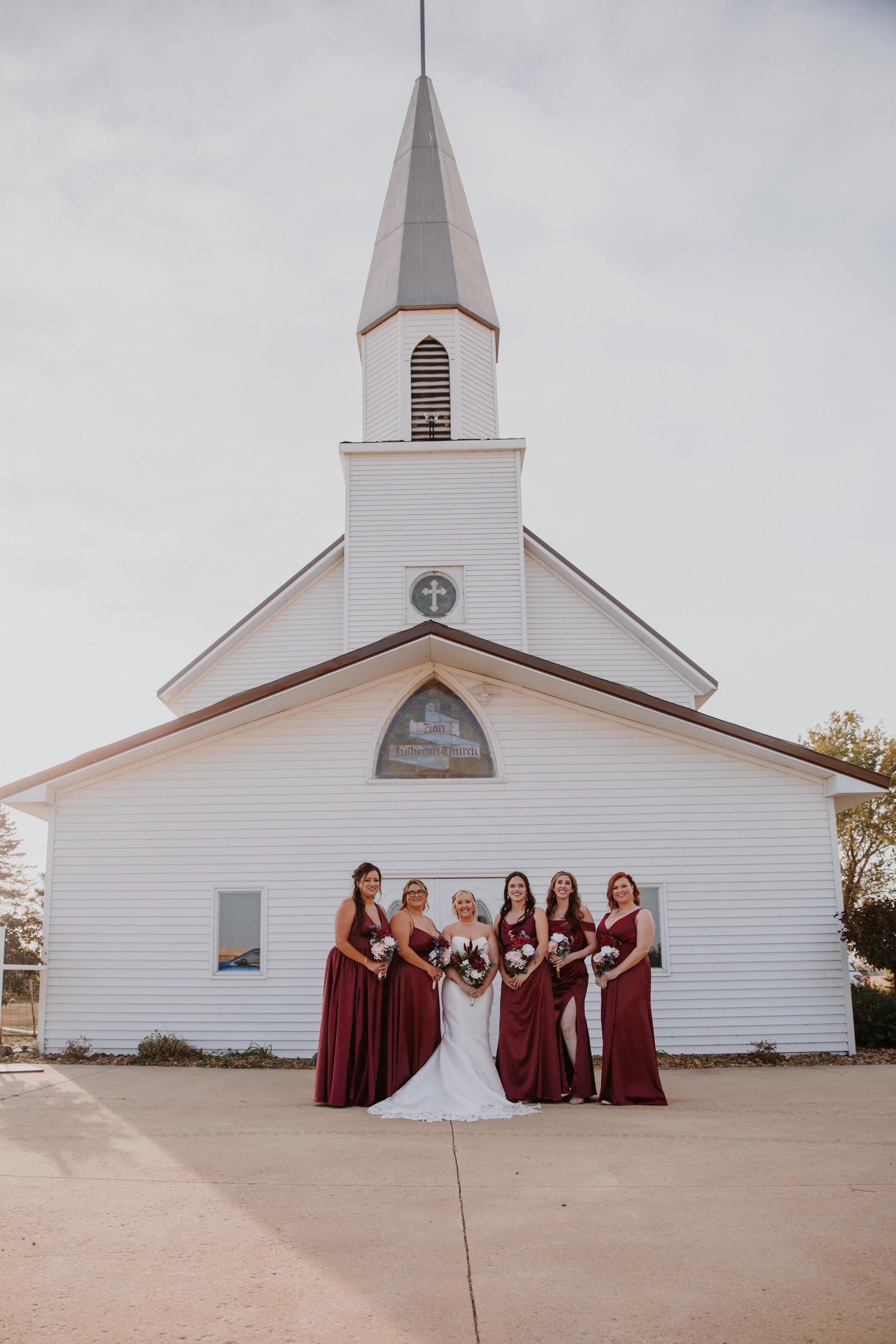 Wedding party in burgundy dresses posed in front of a classic white church exterior.
