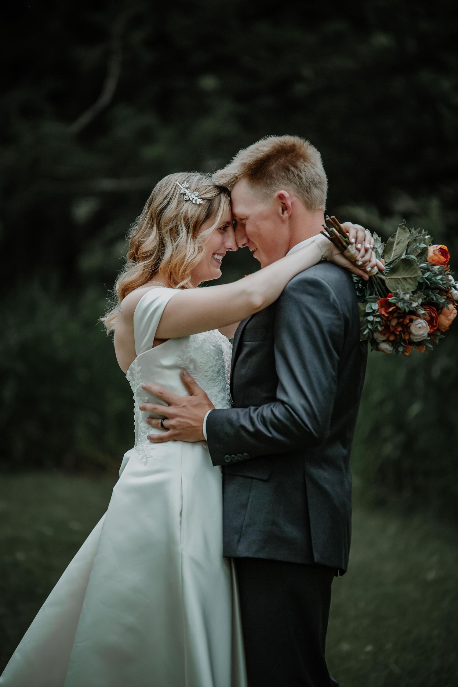 Intimate moment between newlyweds with coral bouquet against dark forest background.