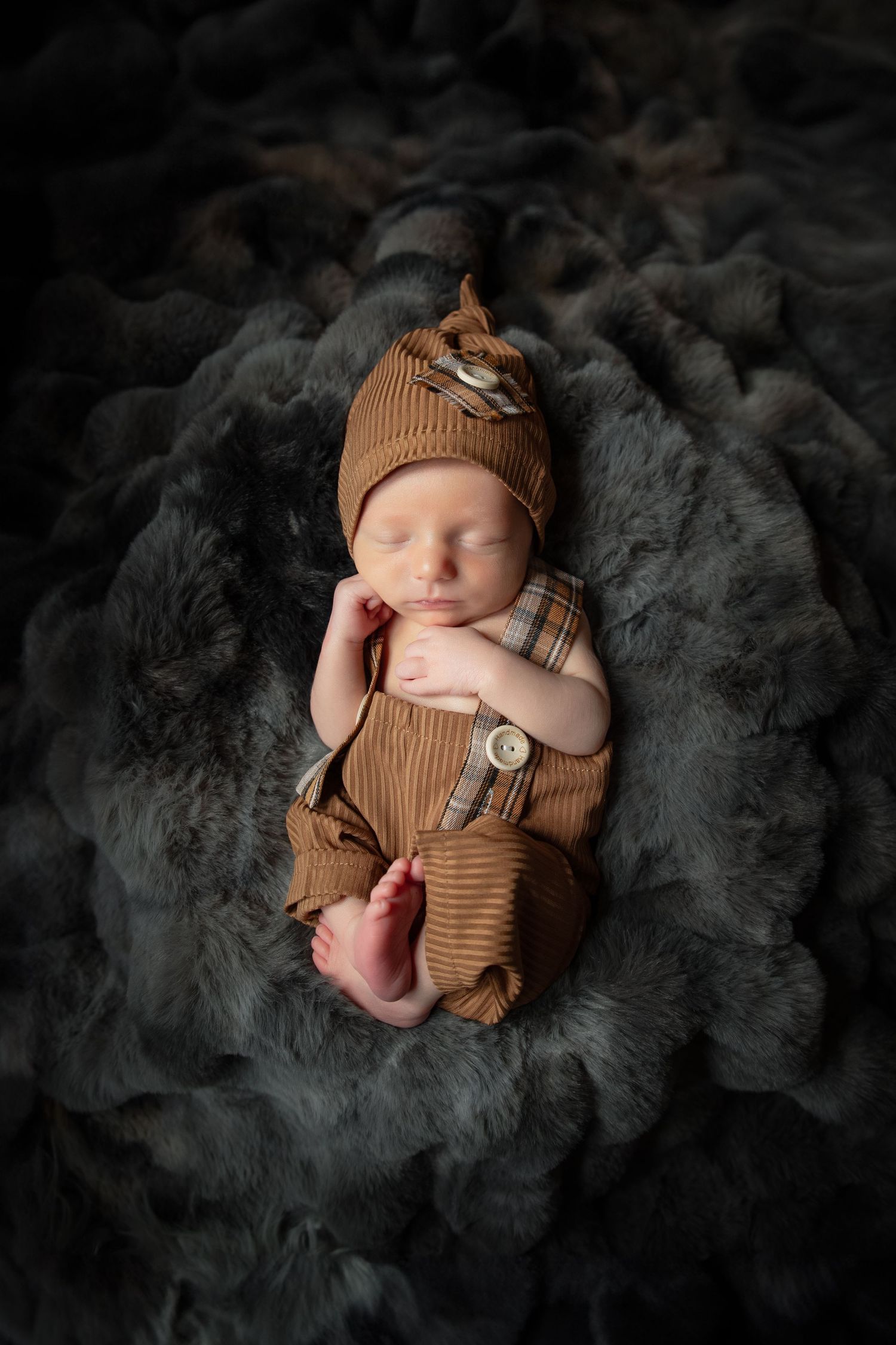Sleeping newborn in brown knit outfit rests peacefully on soft grey textured blanket in dark studio setting.