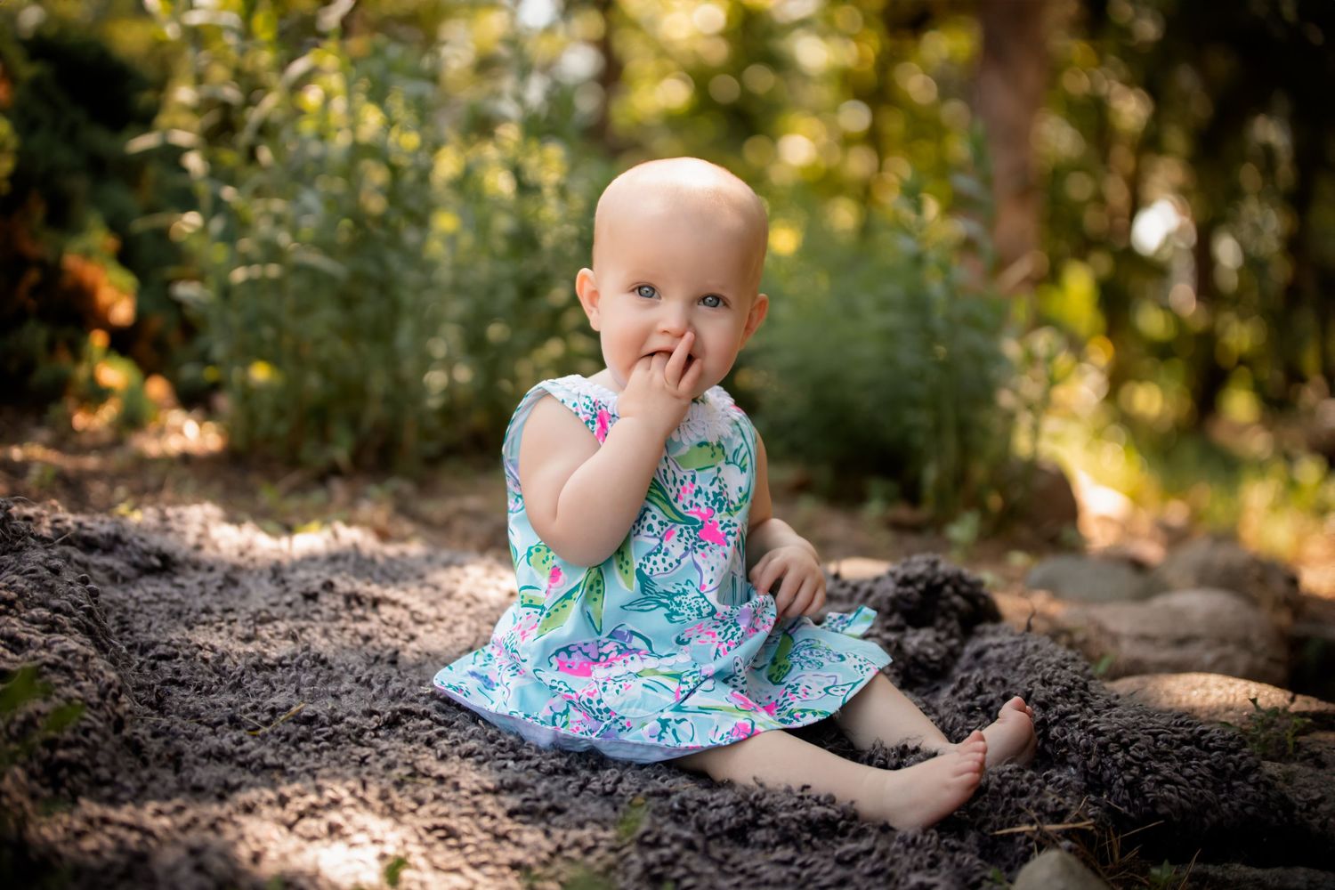 Baby in turquoise dress sits on natural stone path with finger near mouth. Photographer in Sioux Falls, SD