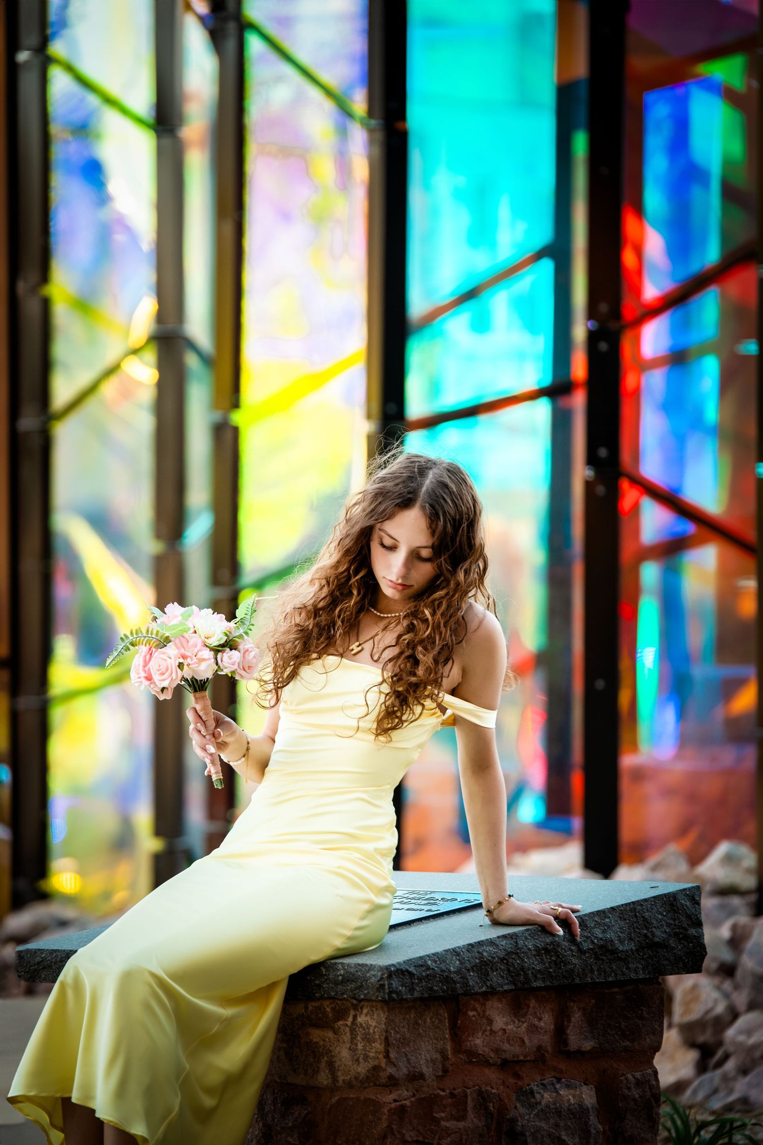 Woman in yellow dress lying on stone wall with colorful stained glass windows creating vibrant backdrop.