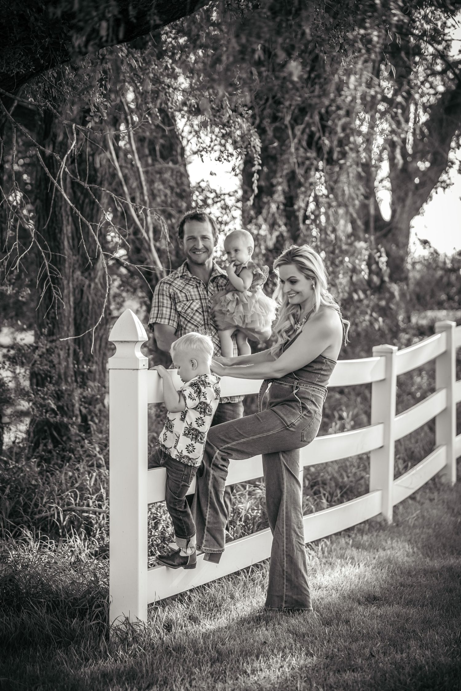 A family poses together by a white fence under tree branches in a black and white photograph.
