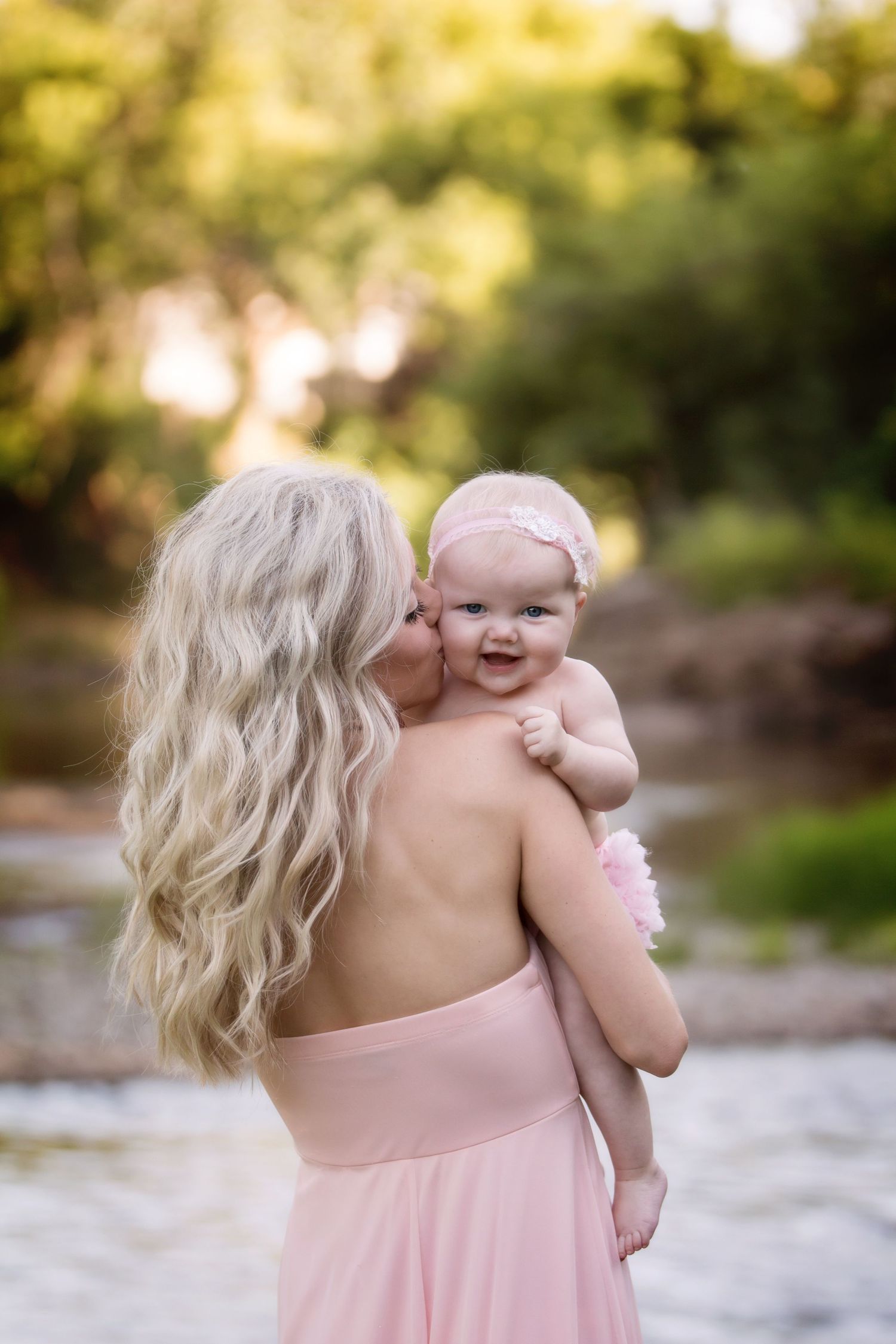 A tender moment between parent and infant is captured near a waterside setting in soft natural lighting.
